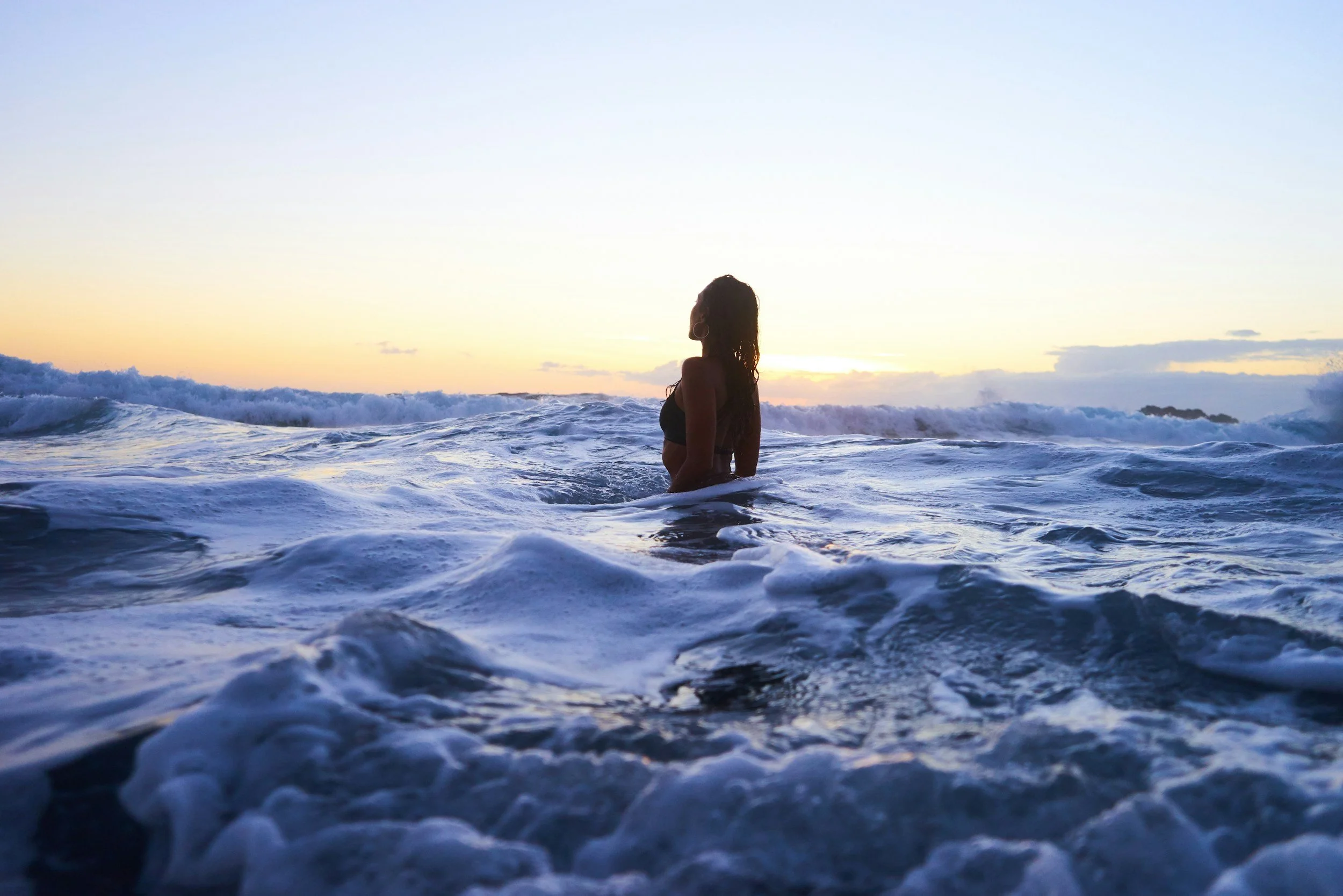 A woman standing in the ocean at sunset, with waves around her and the sky glowing with soft colors.