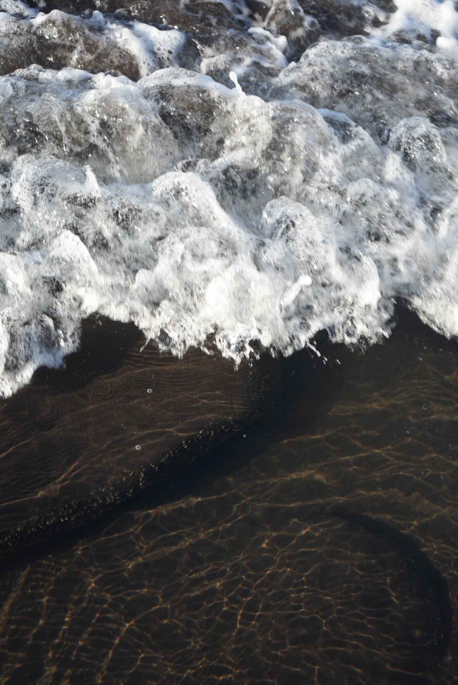 Close-up of ocean waves crashing on the sandy shore, creating white foam with clear water and ripples on the sand.