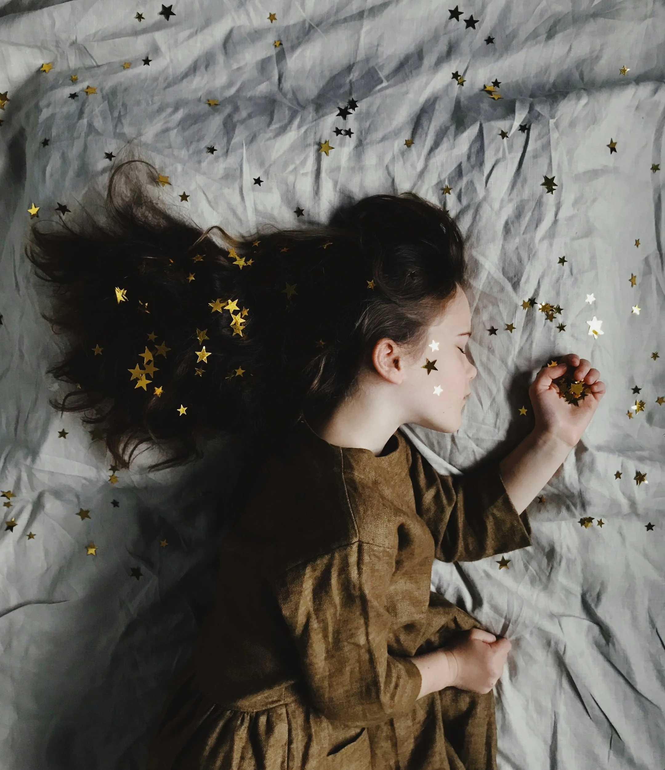 A young girl with dark hair lying on a wrinkled white sheet, surrounded by gold and black star confetti, resting with her eyes closed.
