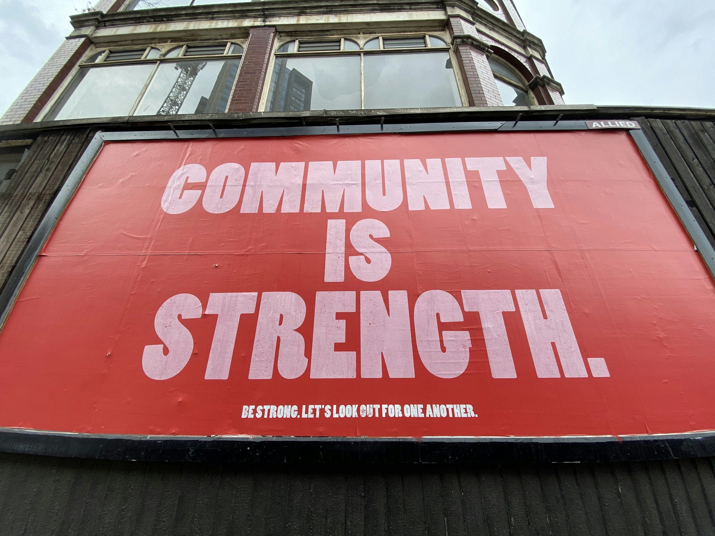 A large red billboard displays the message "Community is Strength" in bold white letters. Below, in smaller white text, it says "Be strong. Let's look out for one another." The billboard is situated on a building with large windows and a brick exterior, and the sky is partly cloudy.
