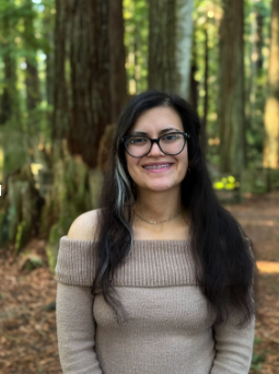 Woman with long dark hair and glasses smiling in a forest setting.