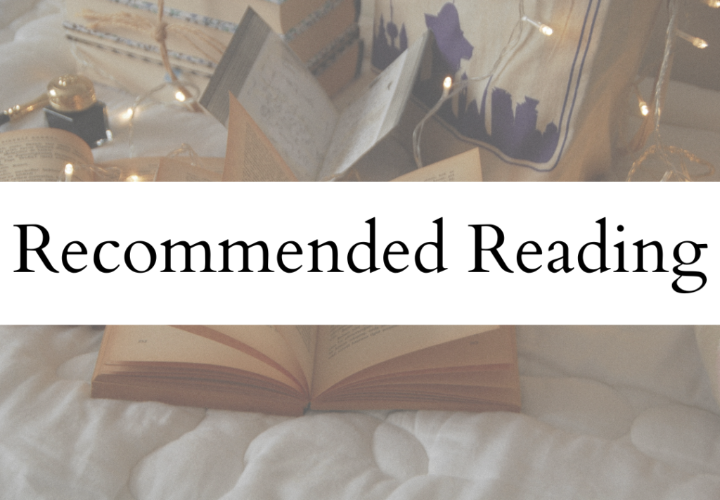 An open book on a bed or blanket with a sign that reads "Recommended Reading" in front of it, surrounded by string lights and other books or decorations.