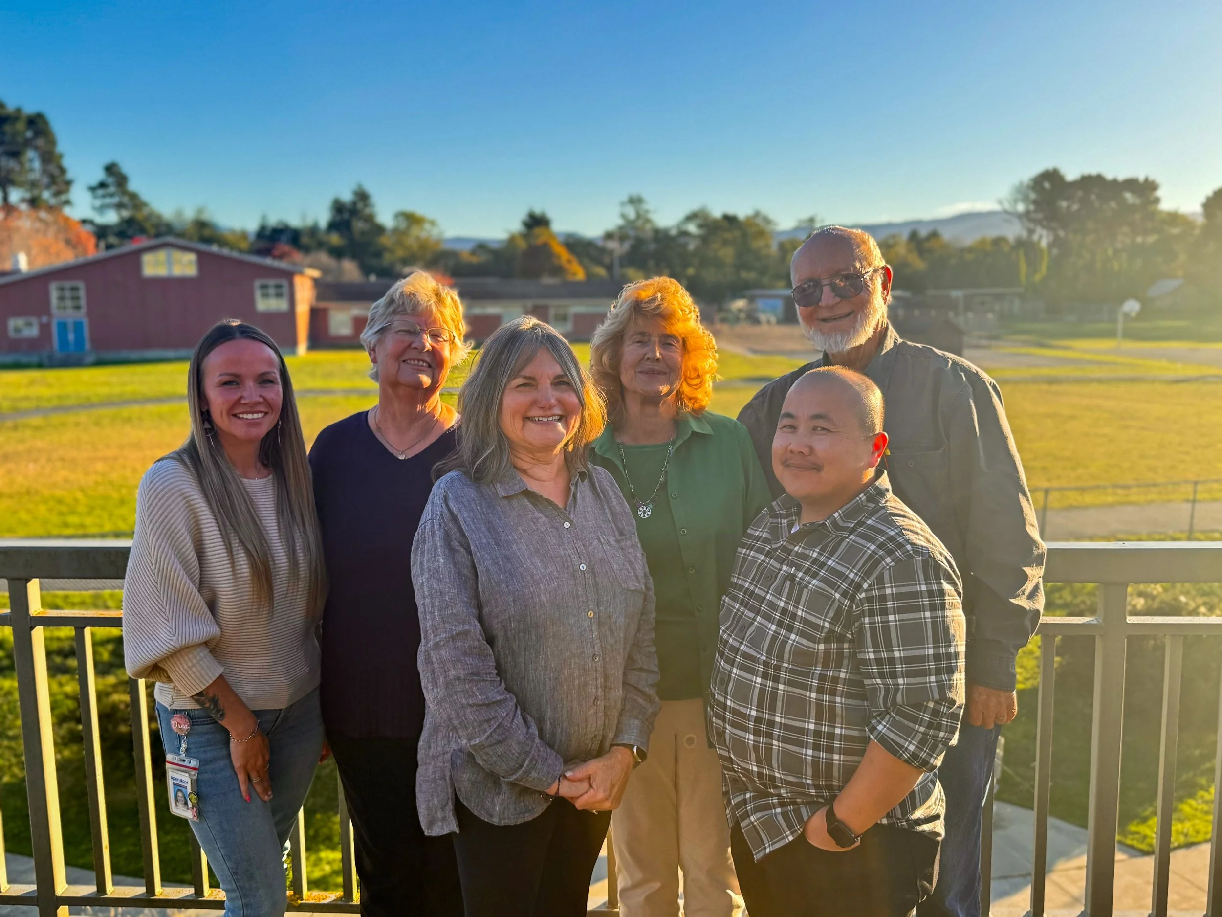 Group of six people(HDVS board members) standing outdoors on a balcony, smiling at sunset with a grassy field, trees, and a red building in the background.