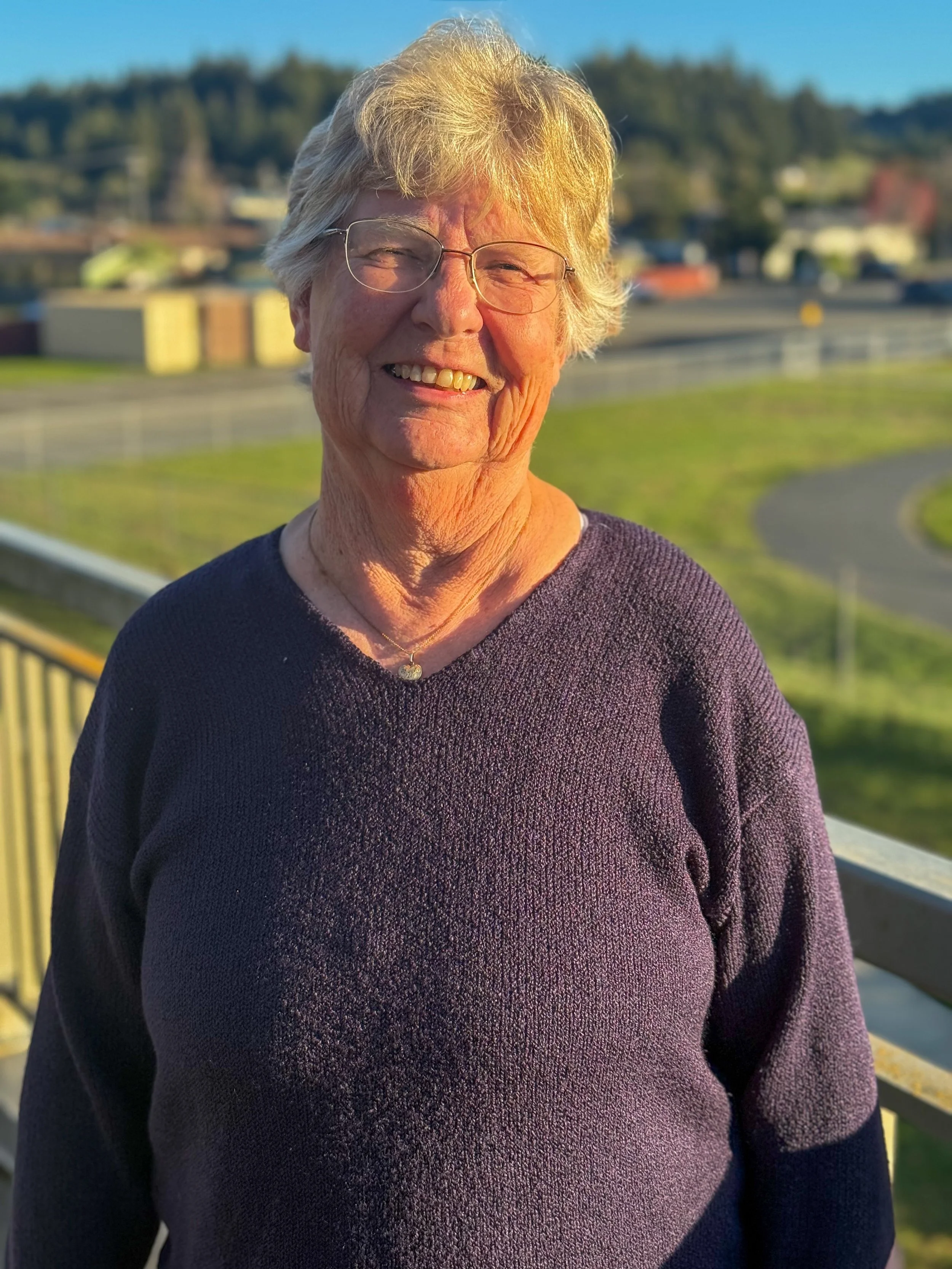 Older woman with white hair and glasses smiling outdoors, wearing a dark sweater, with a green landscape and blue sky in the background.