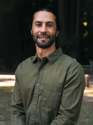 Man with dark hair and beard wearing an olive green button-up shirt standing outdoors in front of trees.