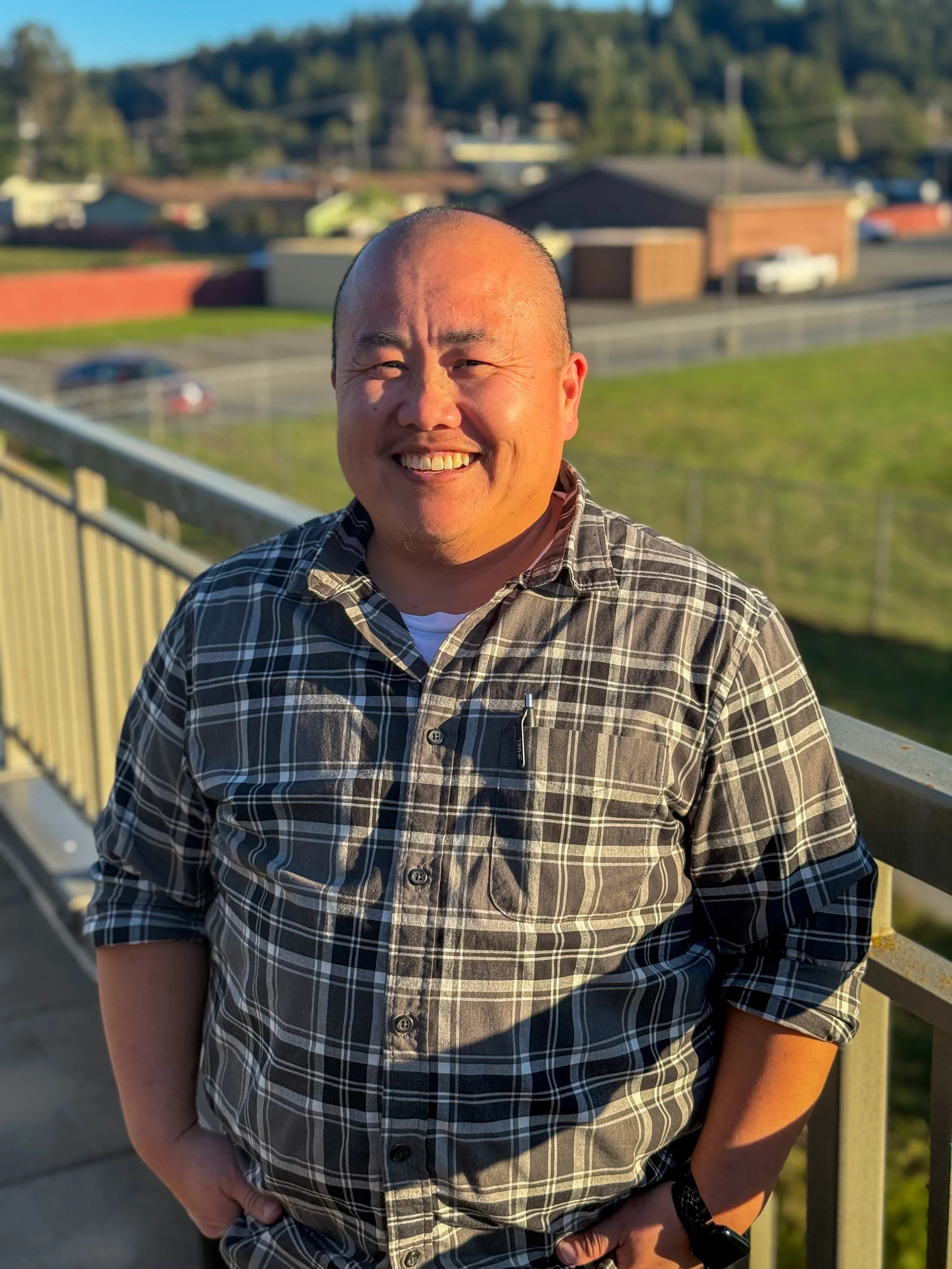 A man with a shaved head smiling outdoors, wearing a black and white plaid shirt with rolled-up sleeves, standing on a balcony or deck with a yellow railing, overlooking a grassy area, a road with cars, and houses in the background during sunset.