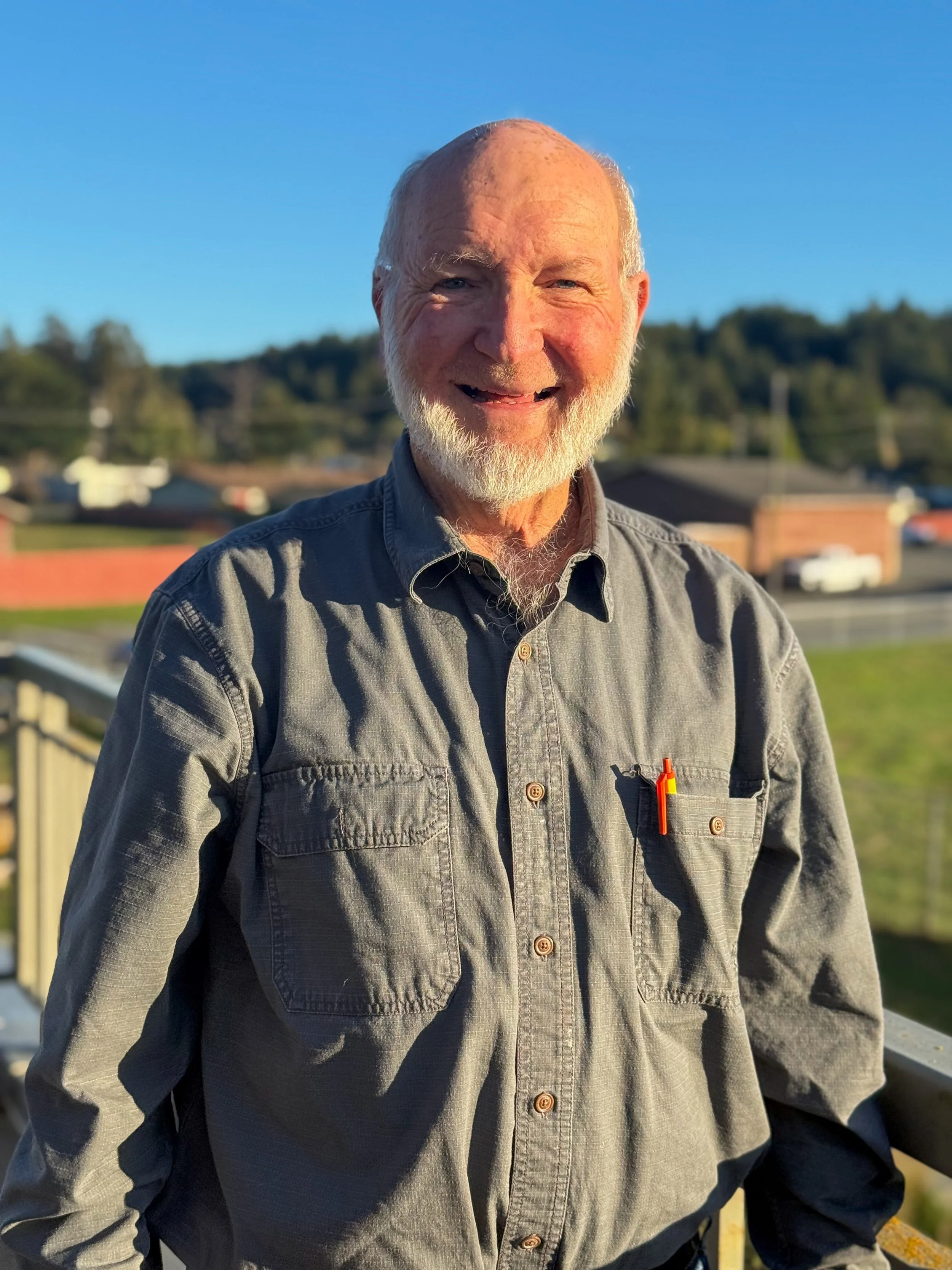An elderly man with a white beard and bald head smiling outdoors on a sunny day, wearing a gray button-up shirt with pens in the pocket, standing on a balcony with a rural neighborhood and trees in the background.