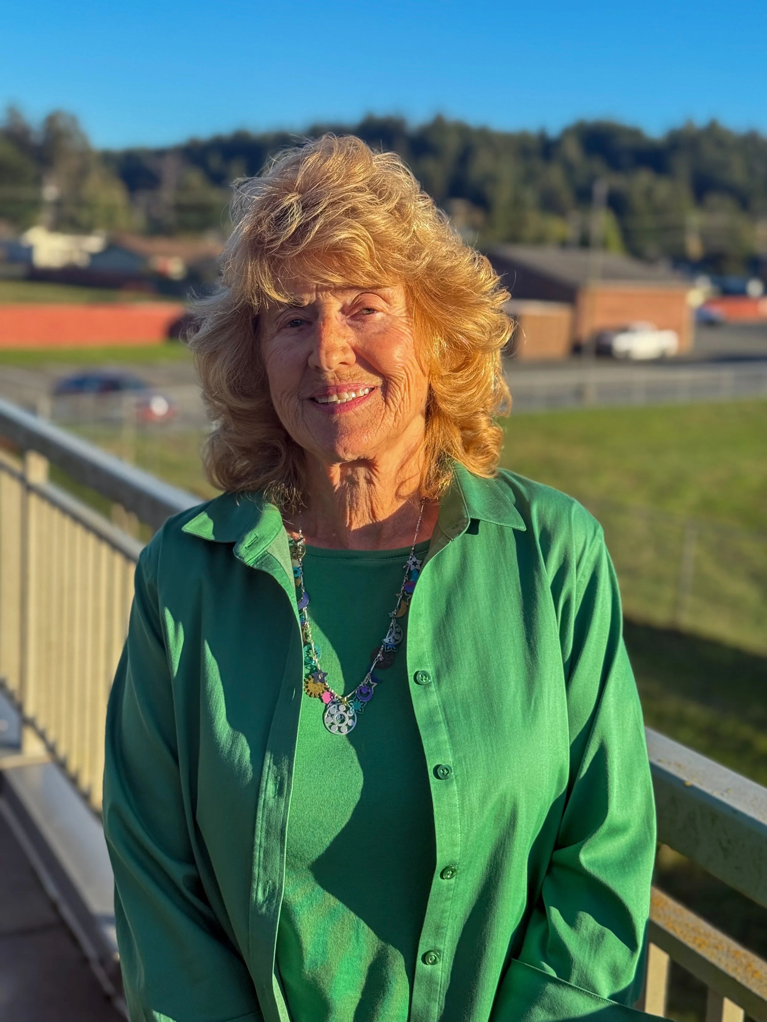 A smiling elderly woman with curly blonde hair stands outdoors on a balcony, wearing a green shirt and necklace, with a blurred background of houses and trees.
