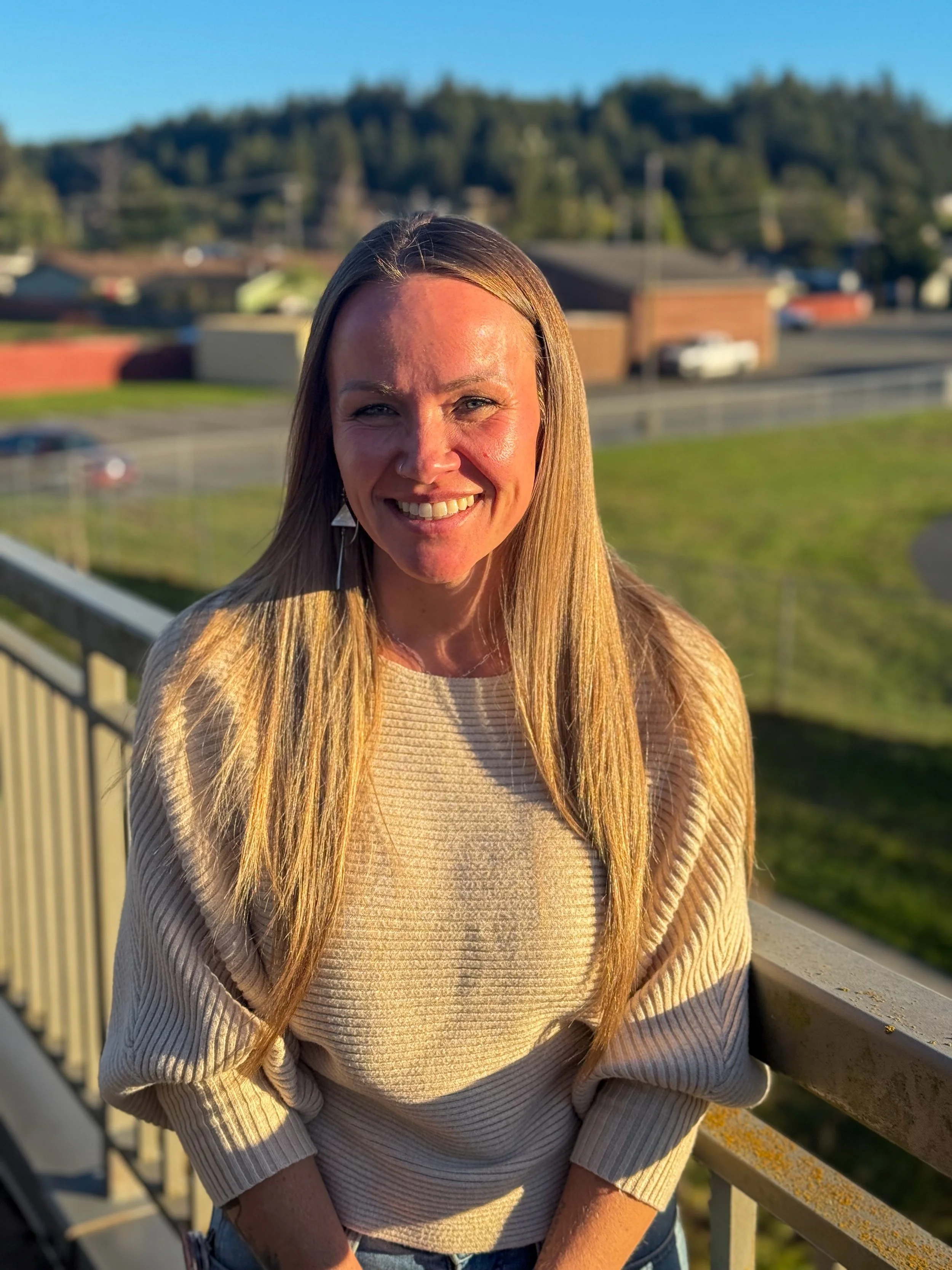 A woman with long blonde hair smiling outdoors on a balcony with a background of trees, buildings, and a parking lot during sunset.