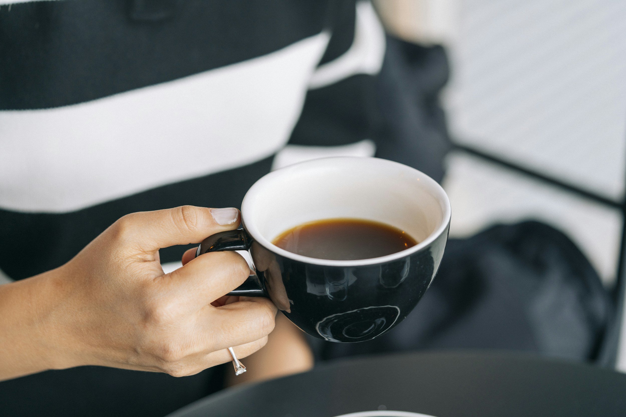 A person holding a black coffee mug with a white interior, filled with black coffee, while sitting.