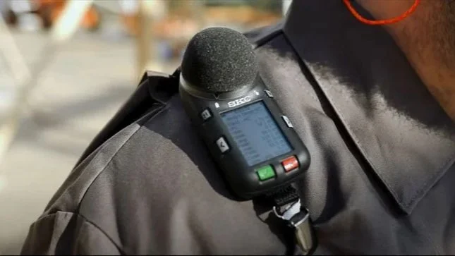 A person wearing a black shirt with a digital sound level meter attached to their shoulder.
