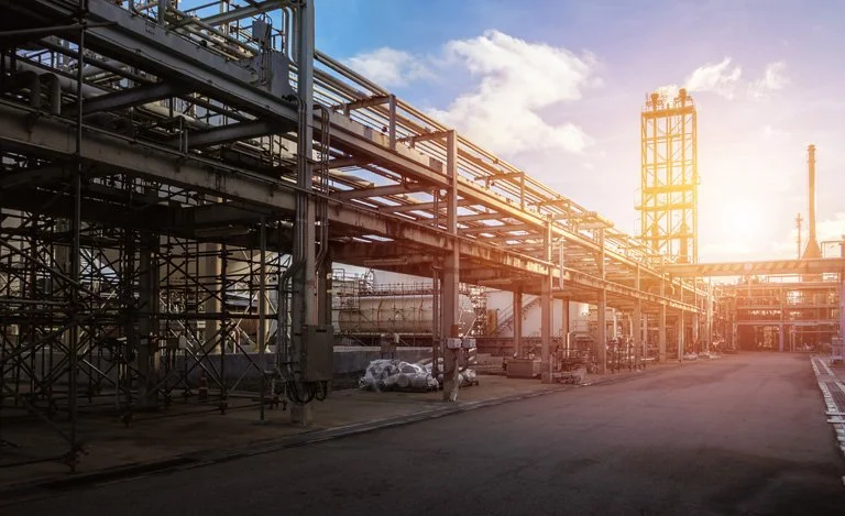 An industrial refinery with metal piping and scaffolding structures, illuminated by sunlight in the late afternoon.