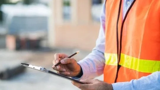 Person wearing a reflective safety vest and holding a clipboard with a pen, standing outdoors.