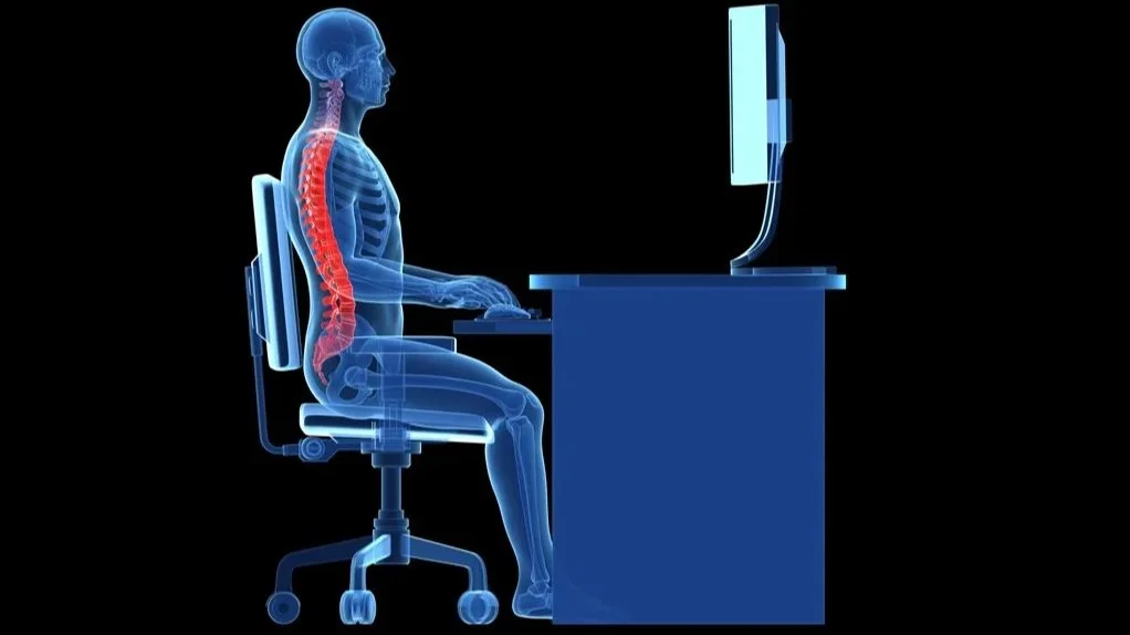 A digital illustration of a human sitting at a desk in front of a computer, with the skeleton and spine visible and highlighted in red.
