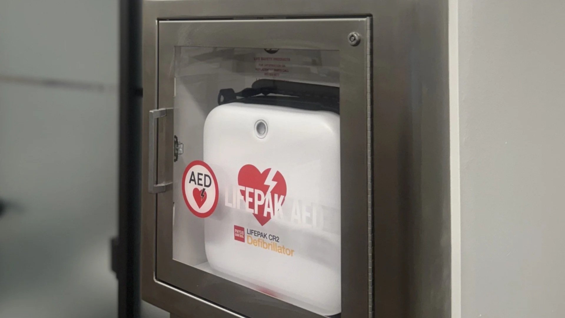 A metal cabinet with a glass front housing an automated external defibrillator (AED) labeled 'LIFEPAK' inside, mounted on a wall in a building.