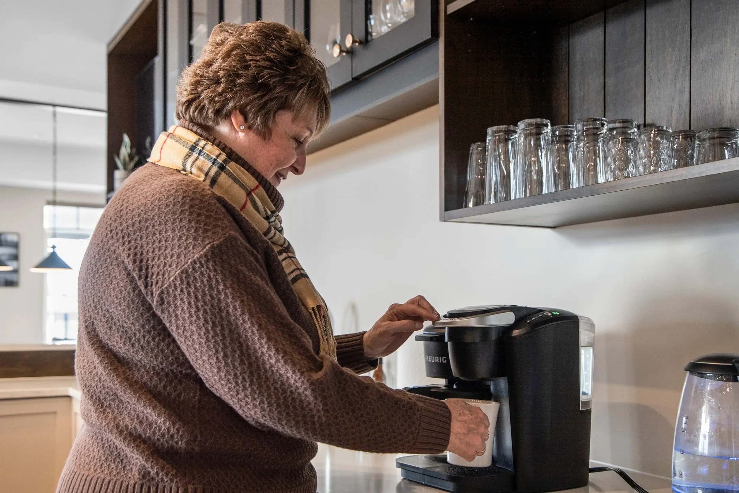 A smiling woman in a brown sweater and a beige Burberry checkered scarf making coffee in a coffee machine in a modern kitchen.