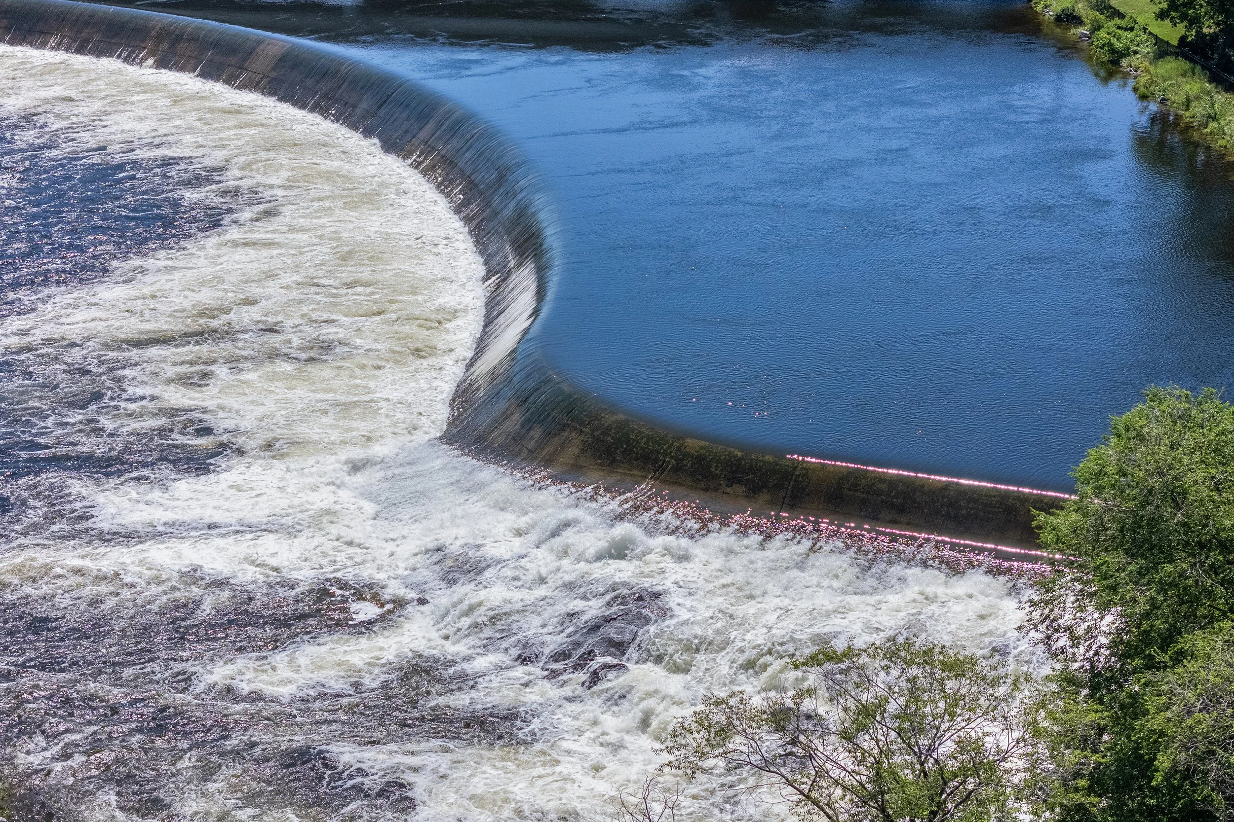 An aerial view of a wide, curved concrete river dam with rushing white water flowing over the spillway
