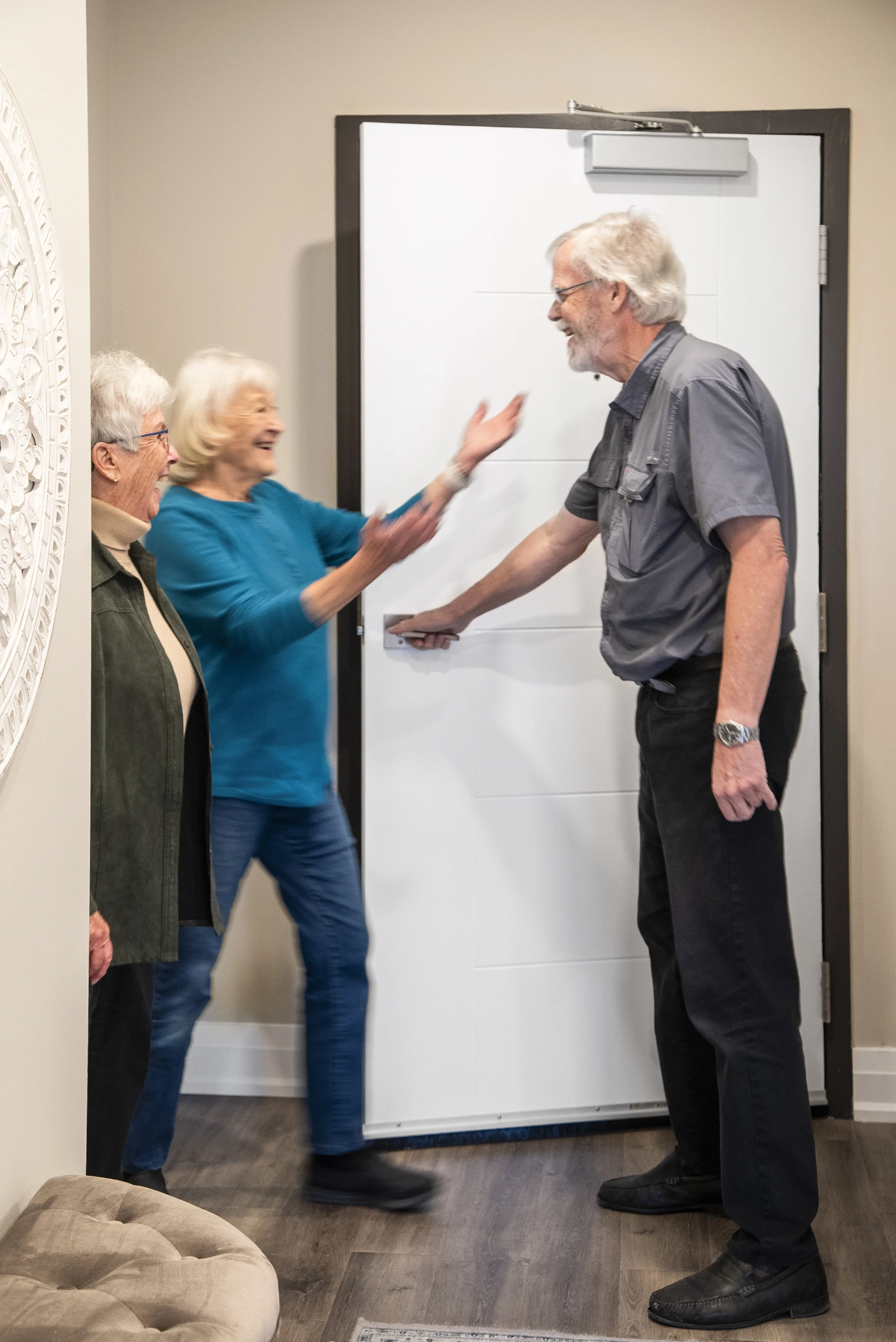 Three elderly people laughing and hugging in front of a white door in a living room.