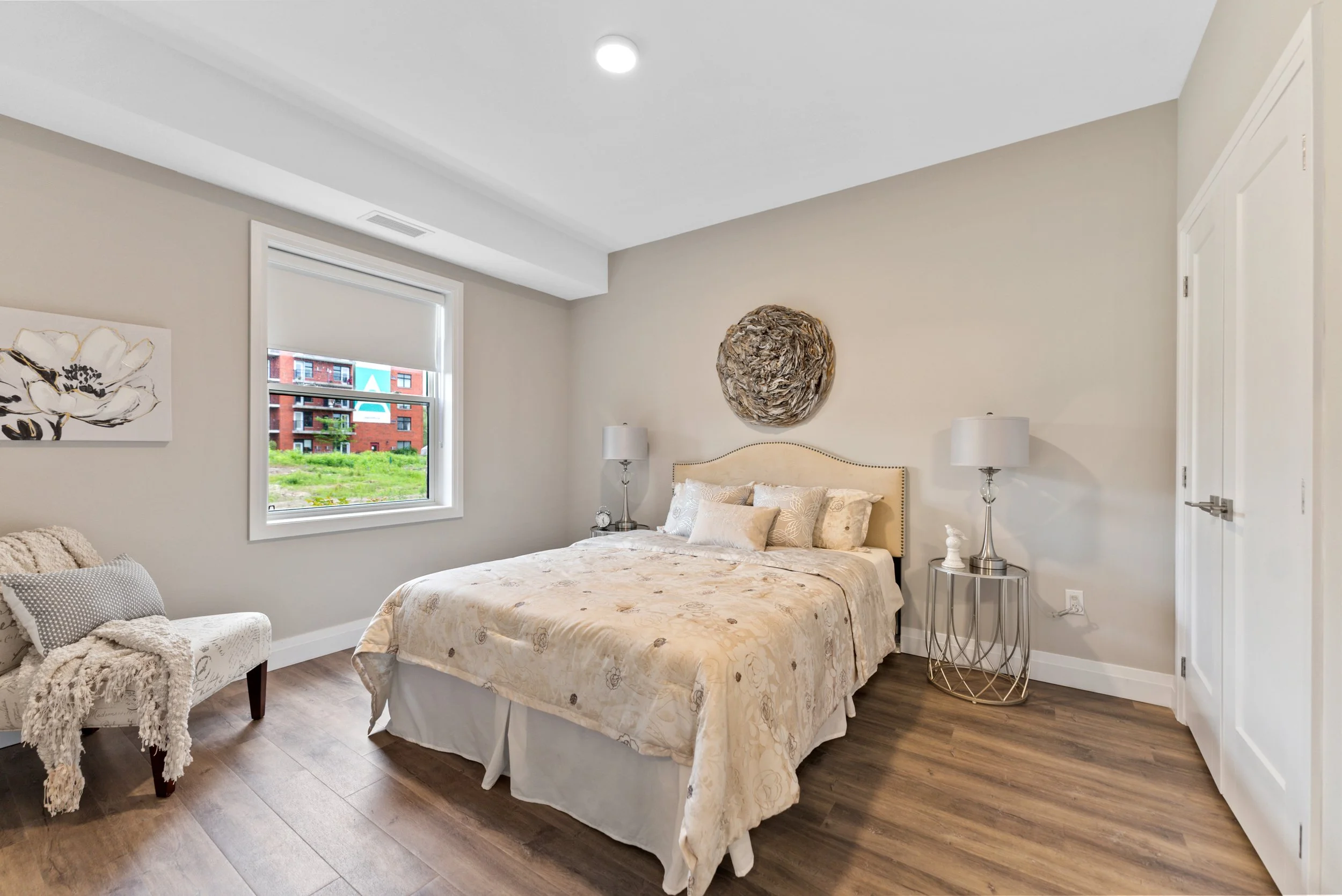 A bedroom with a bed, nightstands with lamps, window with a view of apartments, and wall art.