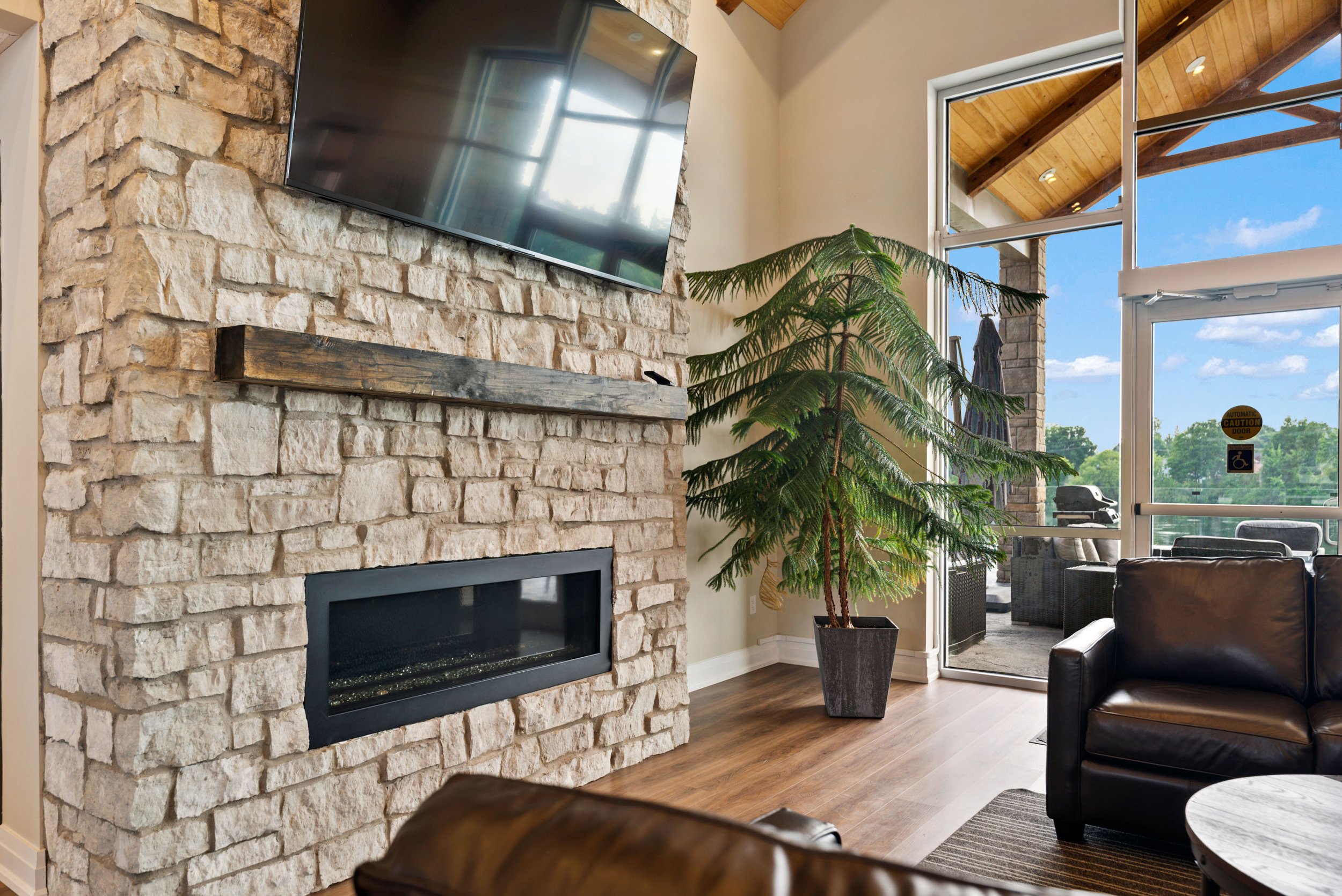 A cozy lounge area featuring a large stone fireplace, a mounted TV, a potted Norfolk Island pine, and leather seating near large glass doors