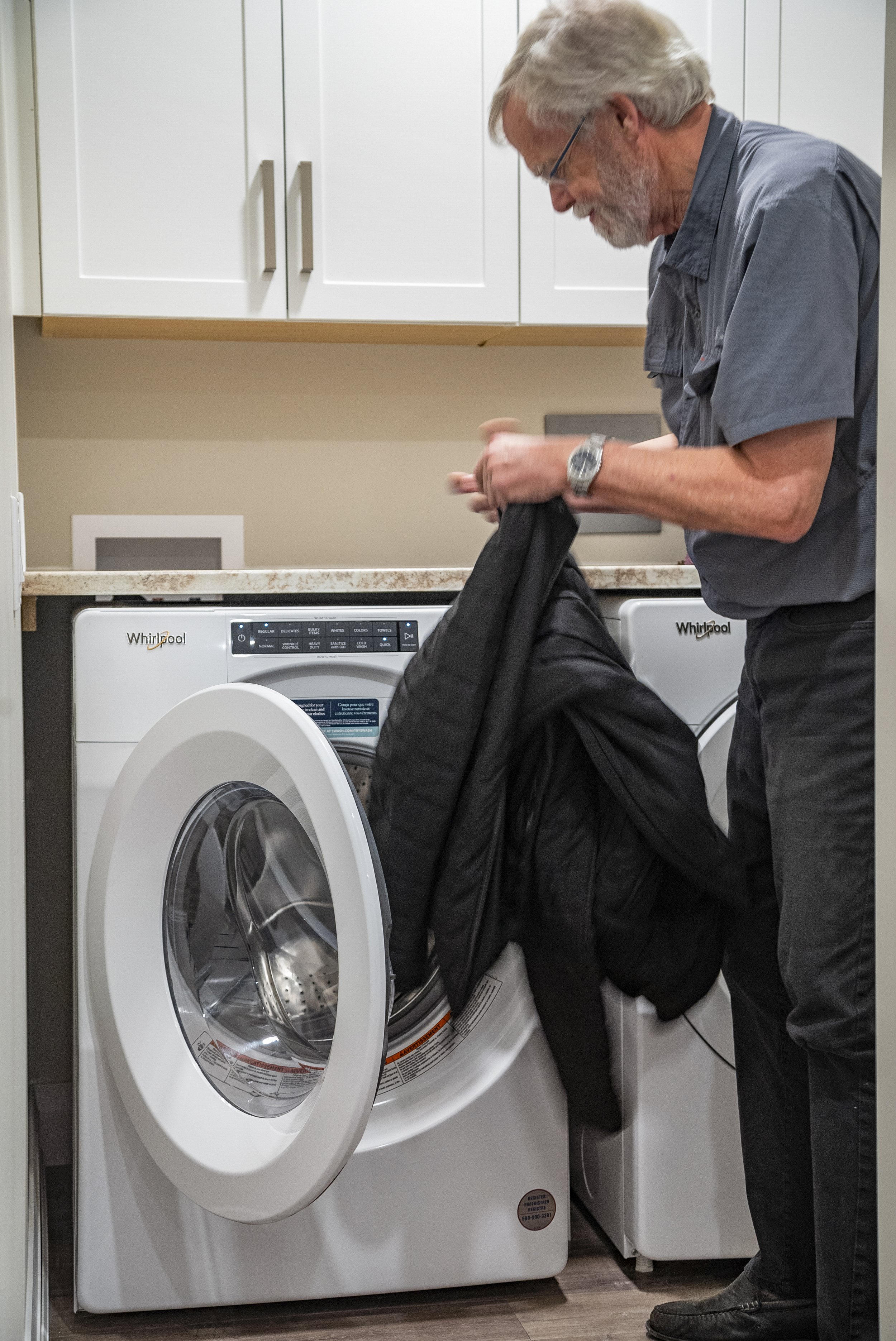 A man loading a black jacket into a front-loading washing machine in a laundry room.