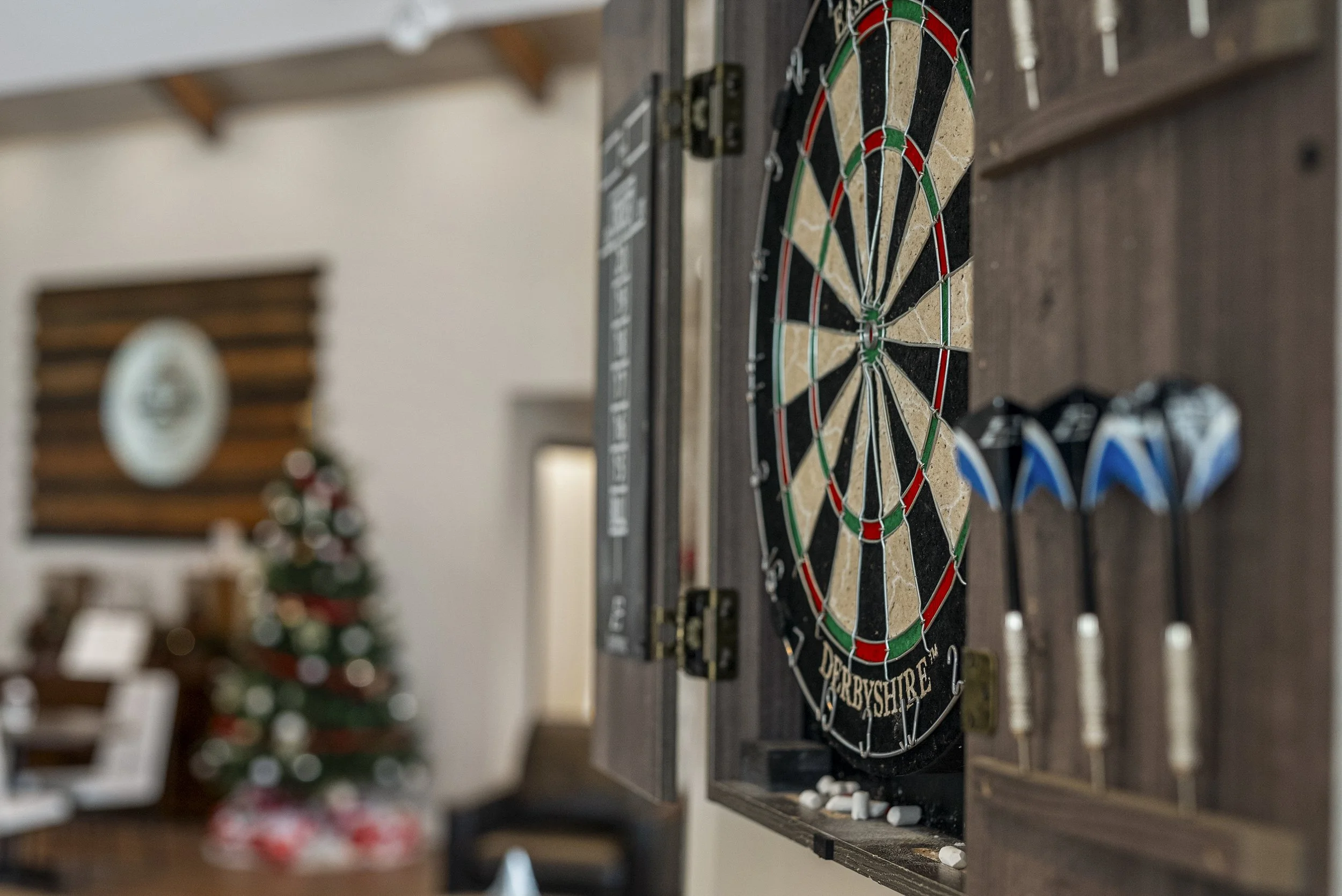 A dartboard with multiple darts stuck in it, mounted on a wall inside a room with wooden decor and a decorated Christmas tree in the background.