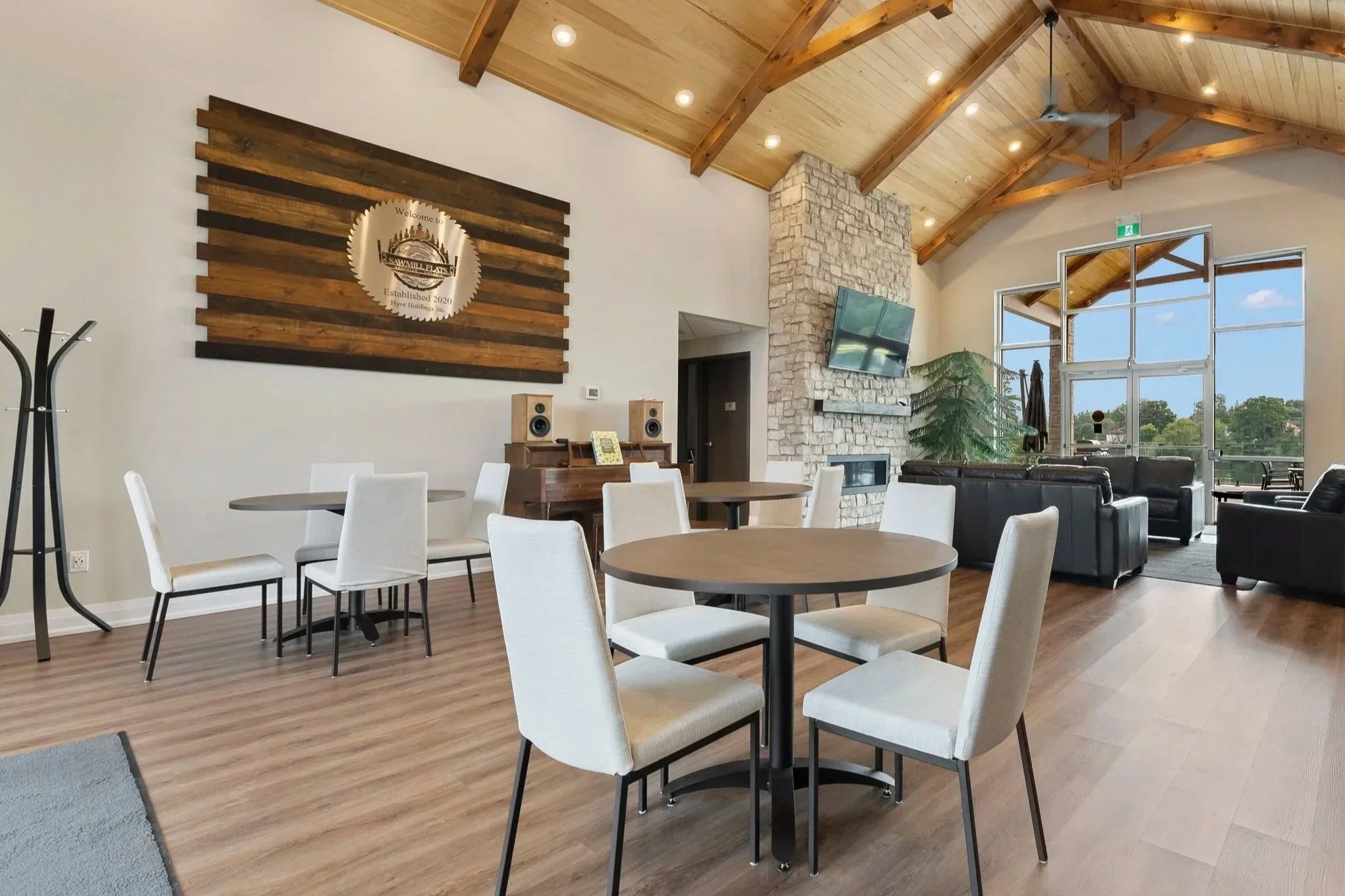 Interior of a living room or lounge area with wooden vaulted ceiling, large windows, a stone fireplace with a TV mounted above, beige walls, and modern furniture including white chairs around round tables, black sofas, a piano, and a coat rack.