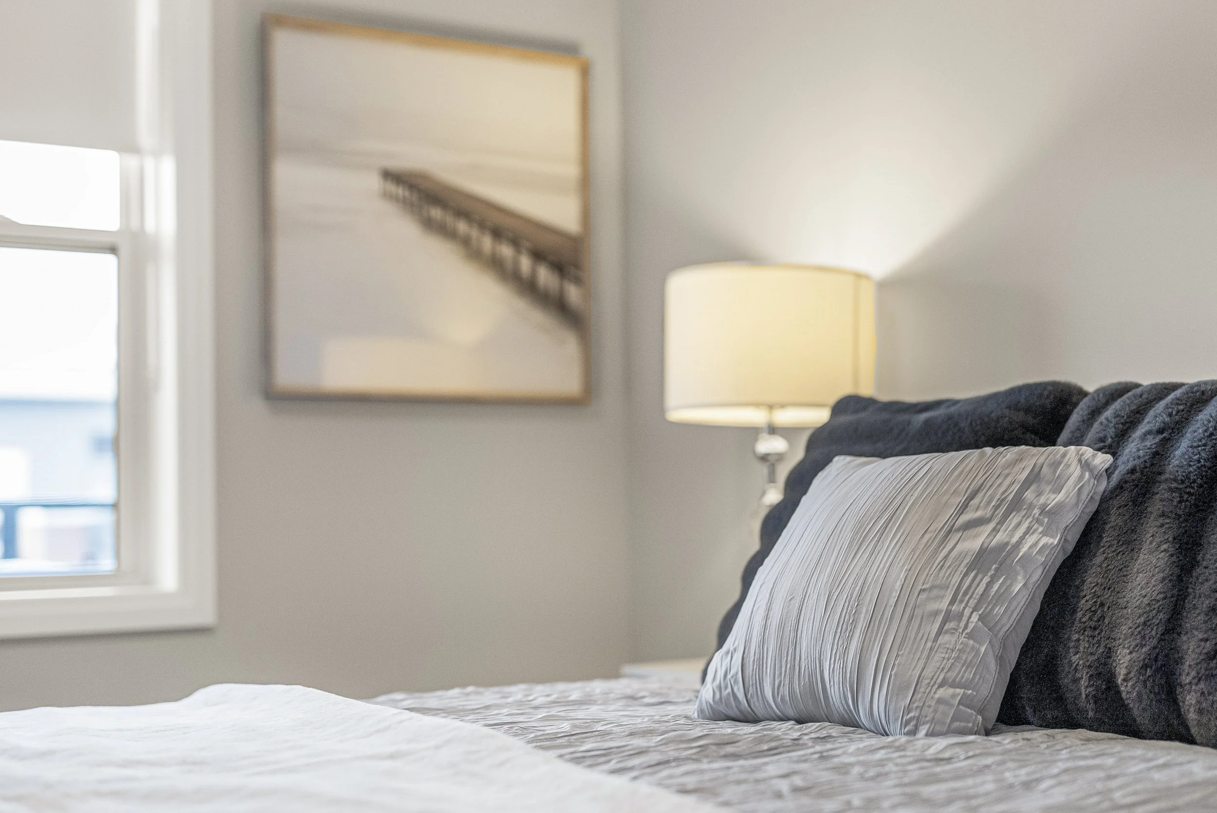 A cozy bedroom with a bed, gray bedding, a matching pillow, a bedside lamp, a window, and a framed picture of a pier on the wall.