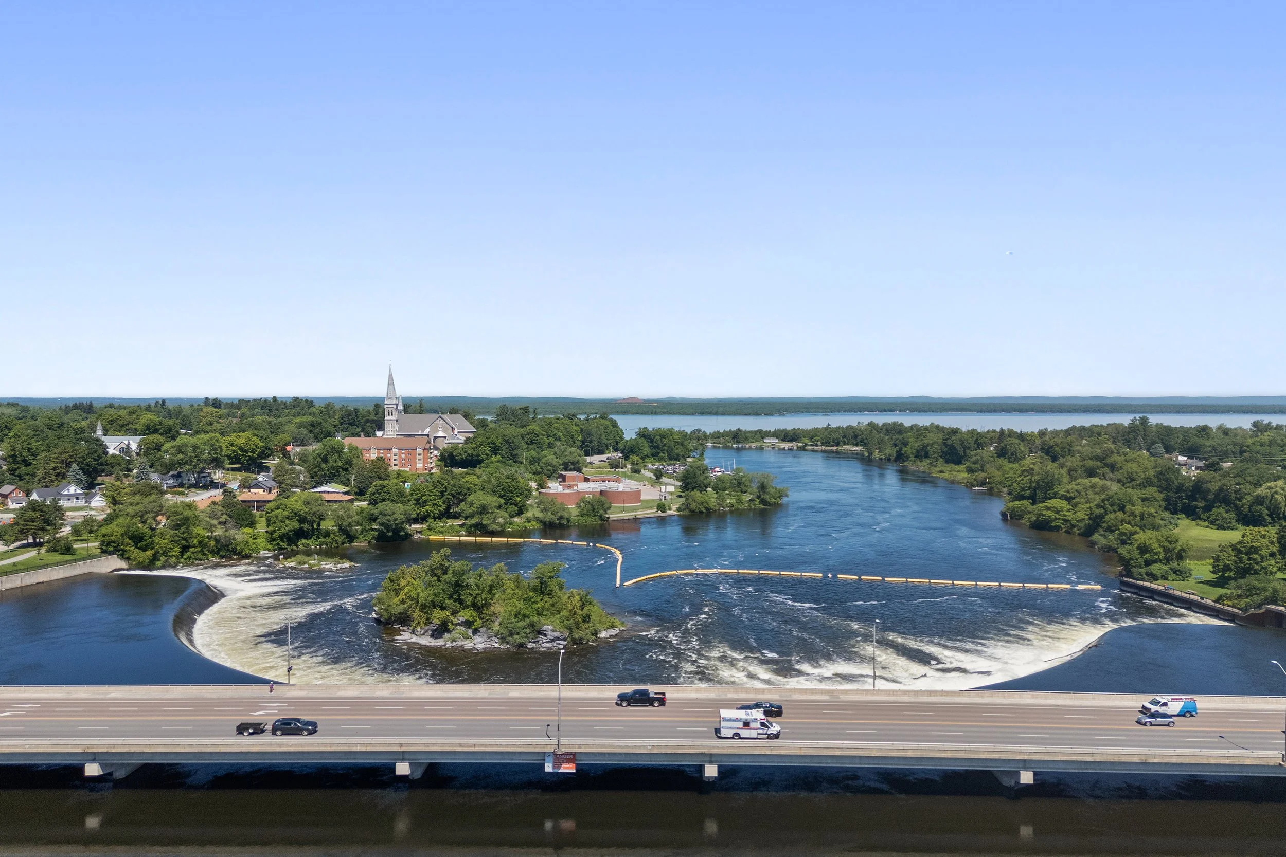 Aerial view of a wide river, dam, and bridge set against a lush green landscape and clear blue sky.