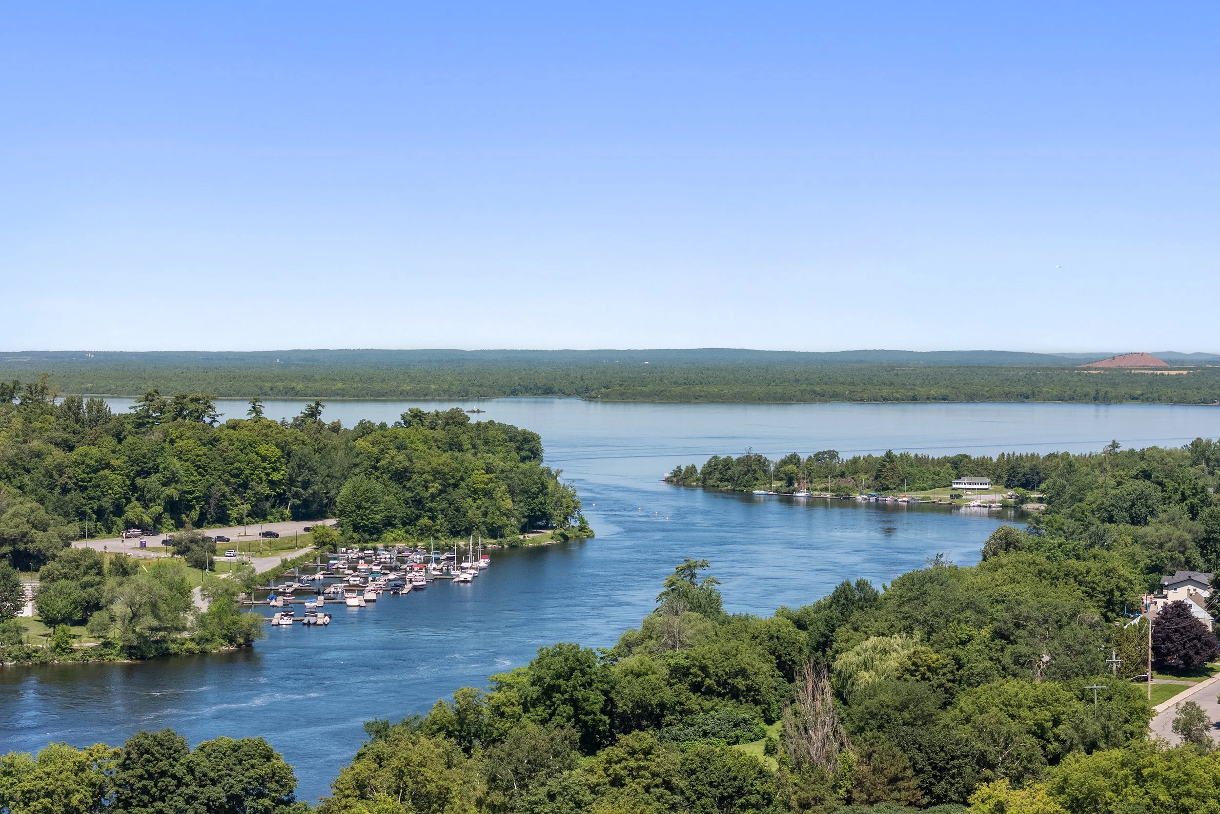 Aerial view of a wide, winding river with a marina full of boats, surrounded by dense green forest and a distant horizon