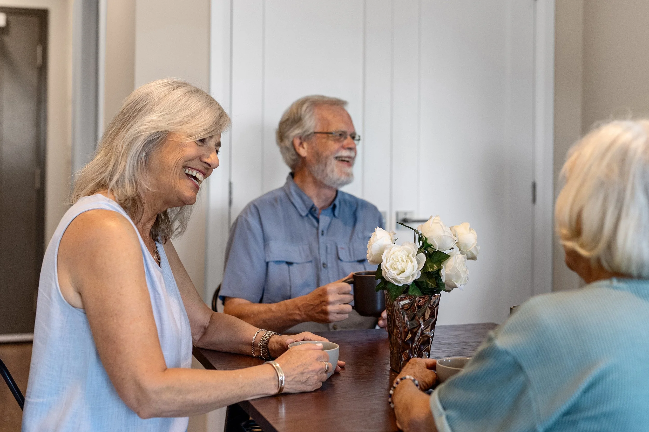 A group of three older adults smiling and chatting over coffee around a dining table decorated with fresh white flowers