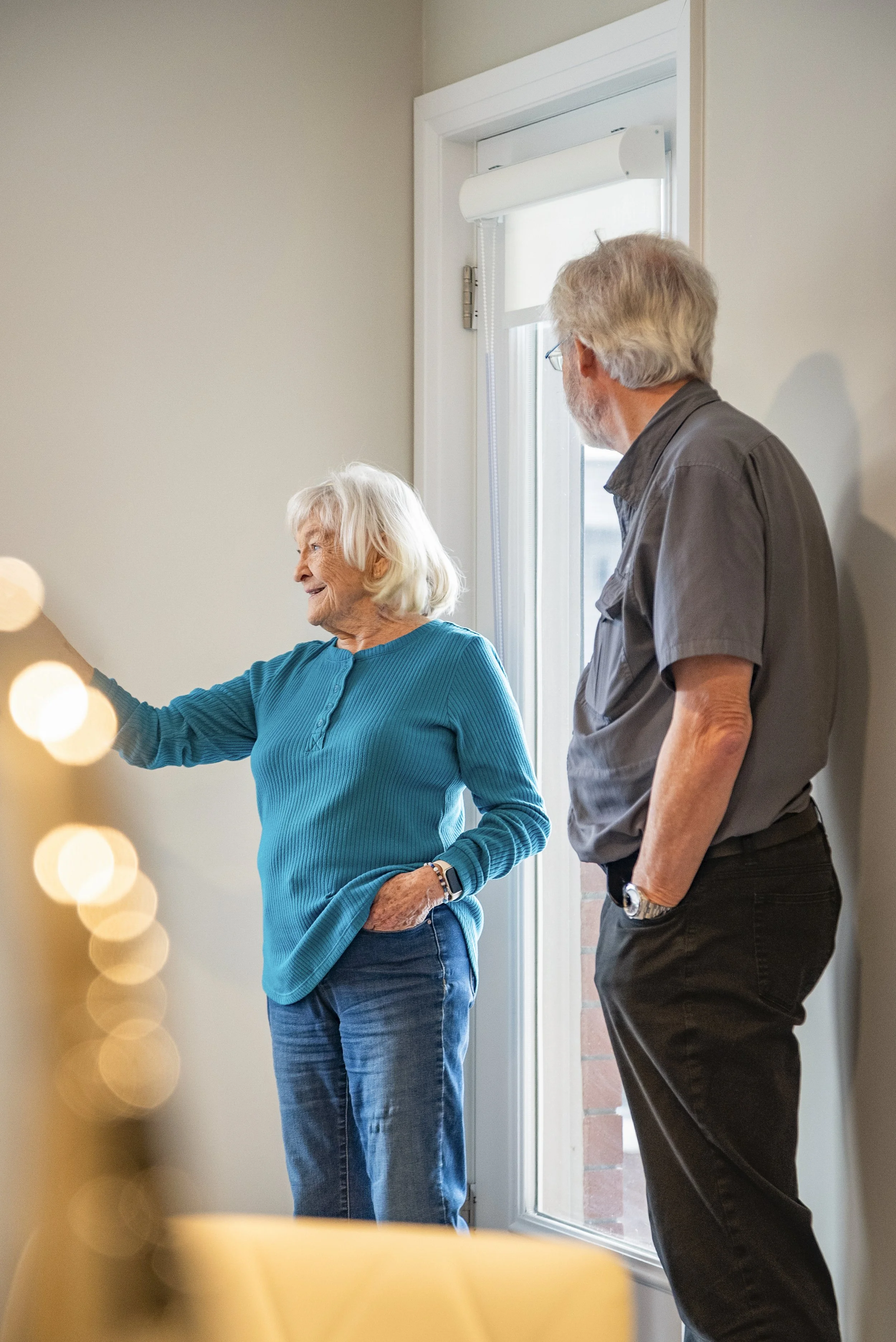An elderly woman with white hair wearing a bright blue sweater and jeans is smiling and gesturing with her right hand while talking to a middle-aged man with gray hair, glasses, a gray shirt, and black pants, standing near a window with white blinds.
