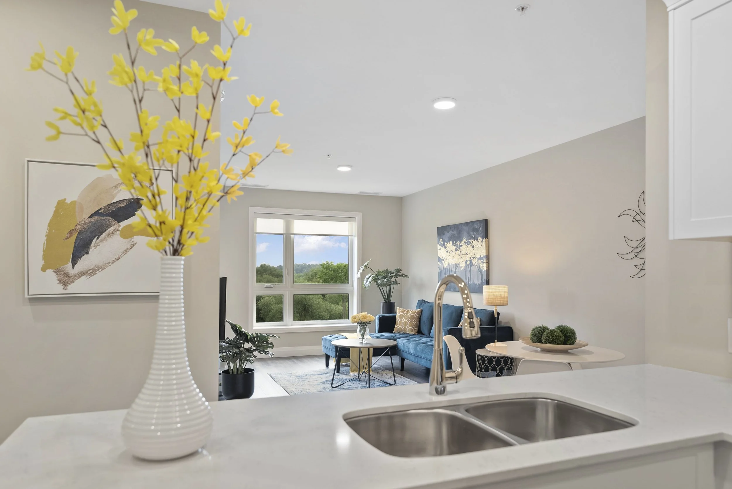 Interior view of a modern living room with a blue sofa, artwork on the walls, a window with trees outside, a small round table with a flower vase, and a lamp. The foreground features a white kitchen counter with a yellow flower arrangement in a white vase and a kitchen sink.