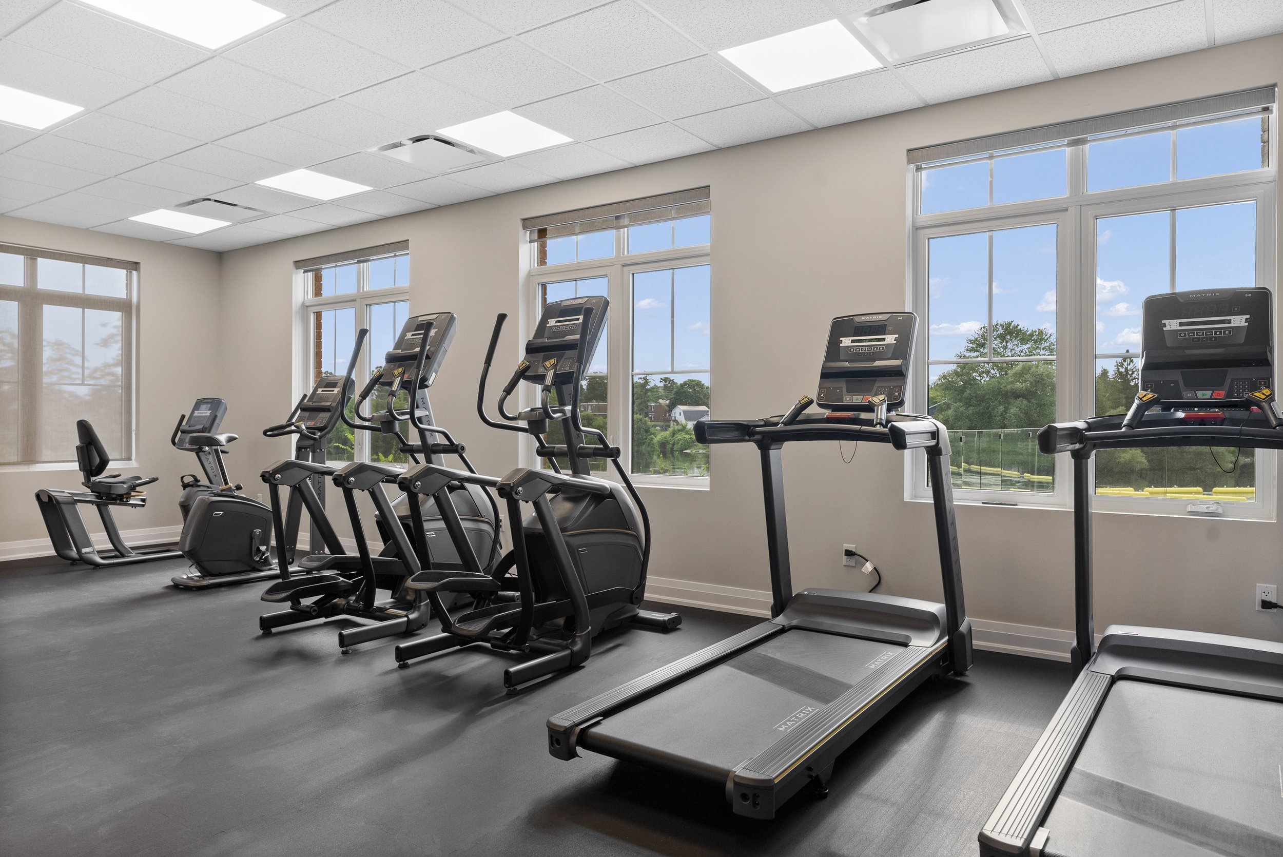 A modern indoor fitness centre featuring treadmills, ellipticals, and stationary bikes facing large windows with a view outdoors  