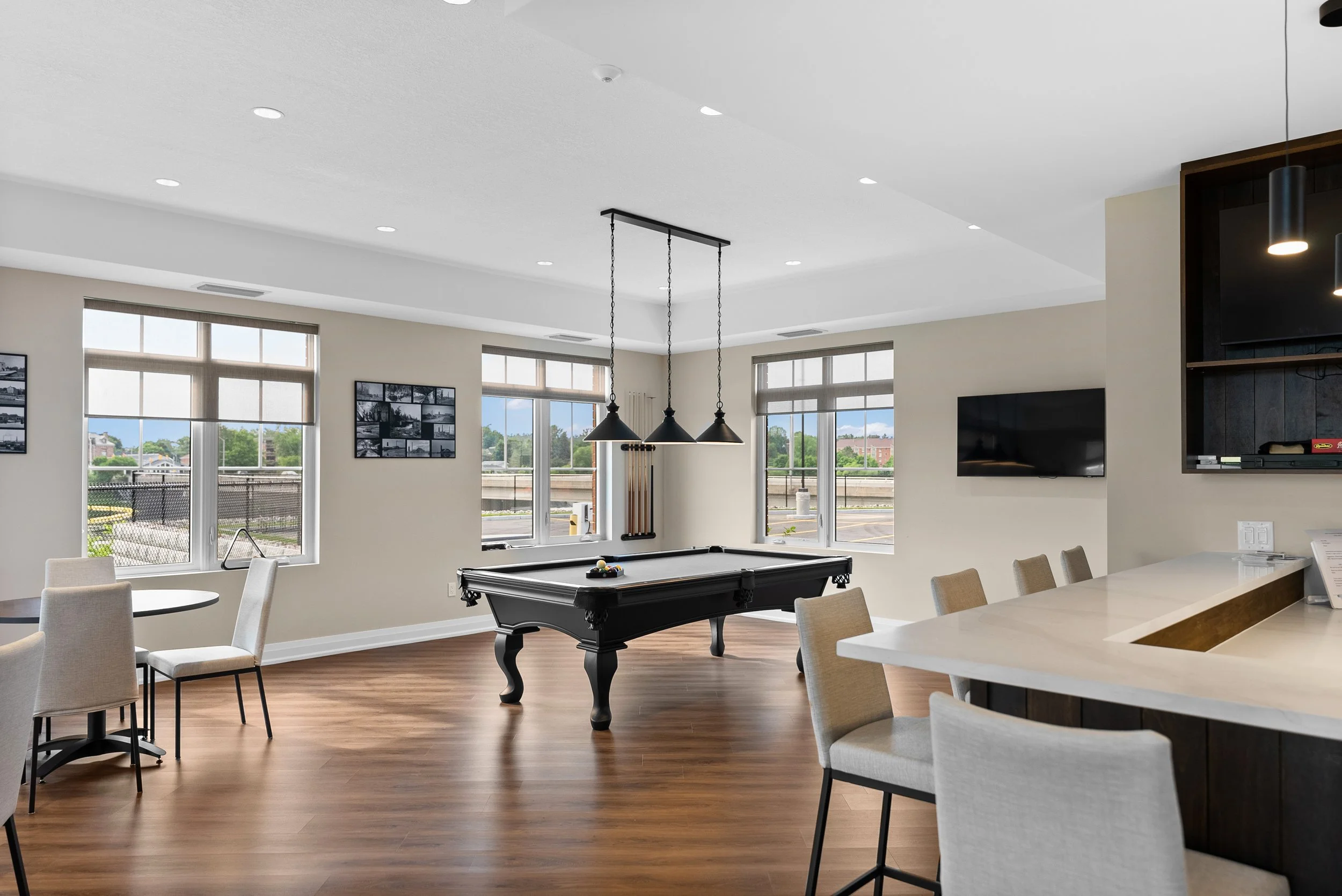 A spacious amenity lounge featuring a central billiards table, pendant lighting, high-top bar seating, and large windows bringing in natural light
