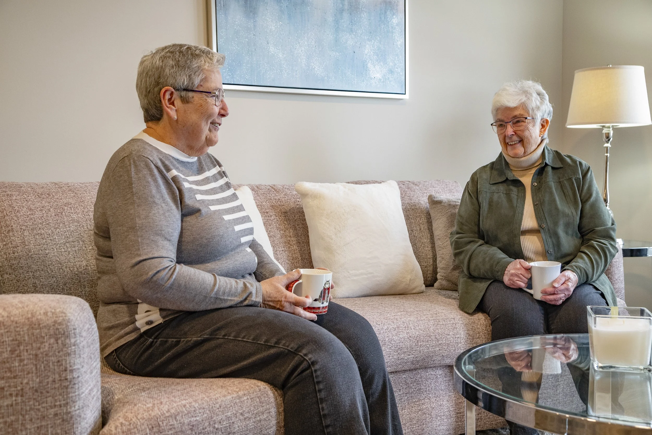 Two elderly women sitting on a beige sofa, smiling and talking, each holding a white mug. A glass-top table with candles is in front of them, with a beige lamp on a side table to the right and a framed picture on the wall behind.