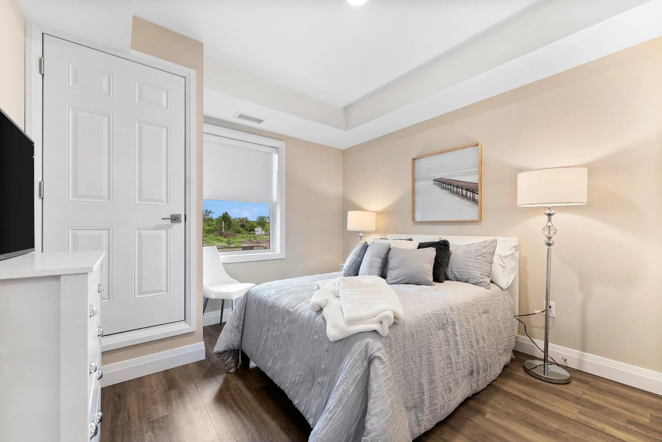 A cozy bedroom with a queen-sized bed, gray bedding, and multiple pillows. There is a nightstand with a lamp on the right and a window with a white blind showing greenery outside. A framed picture of a pier hangs above the bed, and the room has wood flooring and white walls.