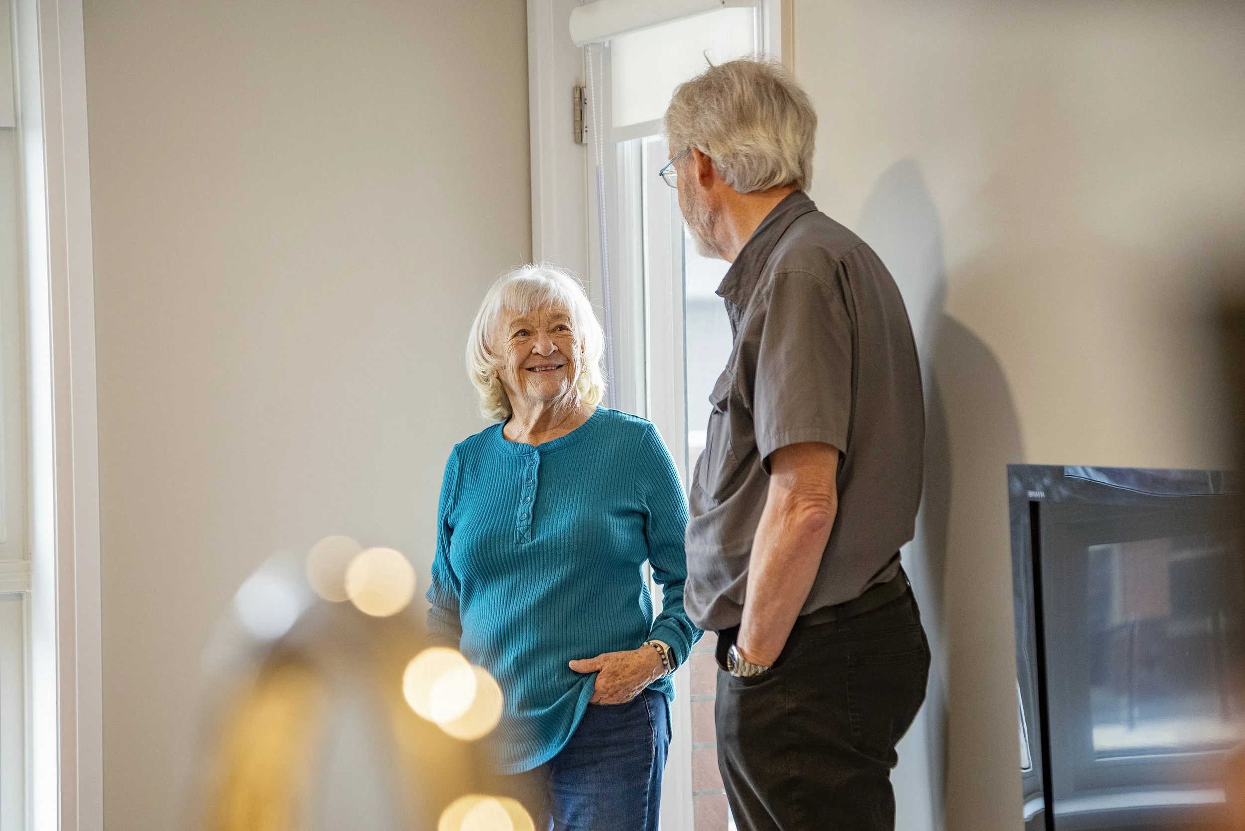 An elderly woman and a middle-aged man are standing and talking inside near a door with a window. The woman is smiling and wearing a bright blue top, while the man is wearing glasses and a gray shirt with dark pants.