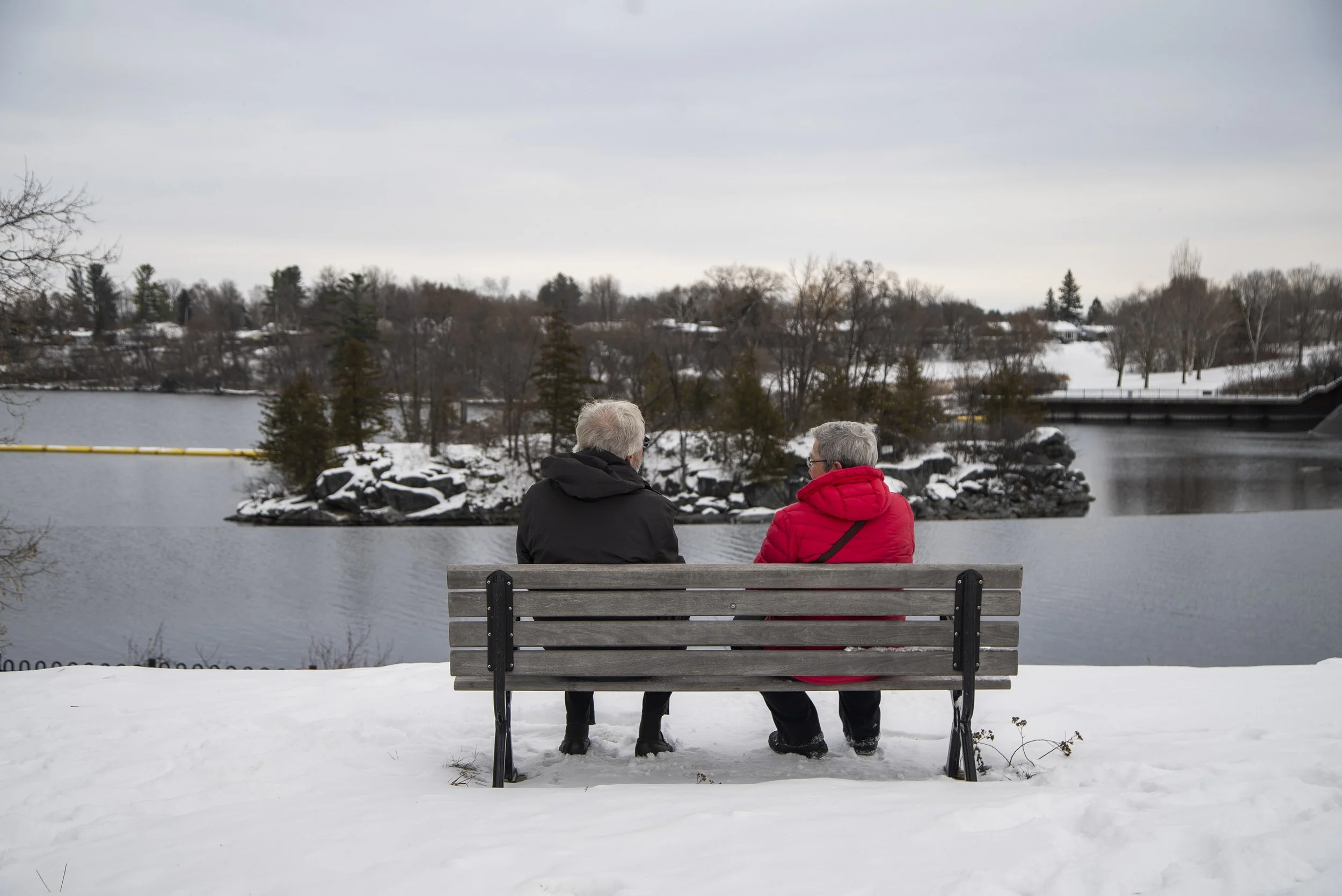 Two elderly people sitting on a park bench by a winter lake, engaged in conversation, with snow-covered ground and leafless trees in the background.