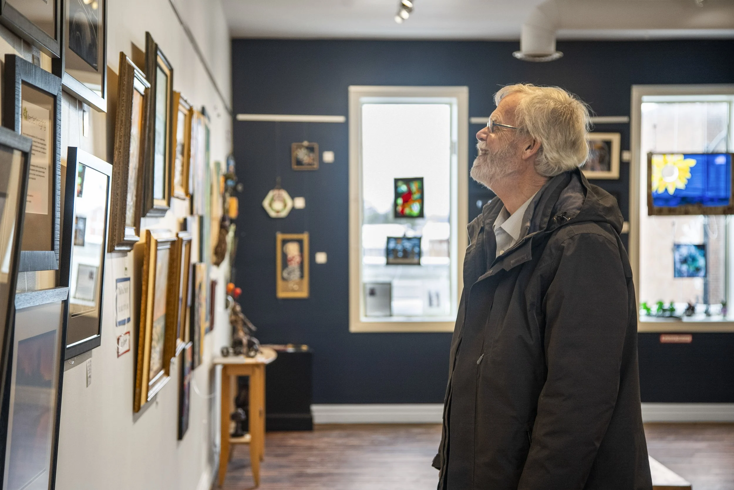 An elderly man with gray hair and glasses looking at framed pictures displayed on a gallery wall inside an Arnprior art gallery.