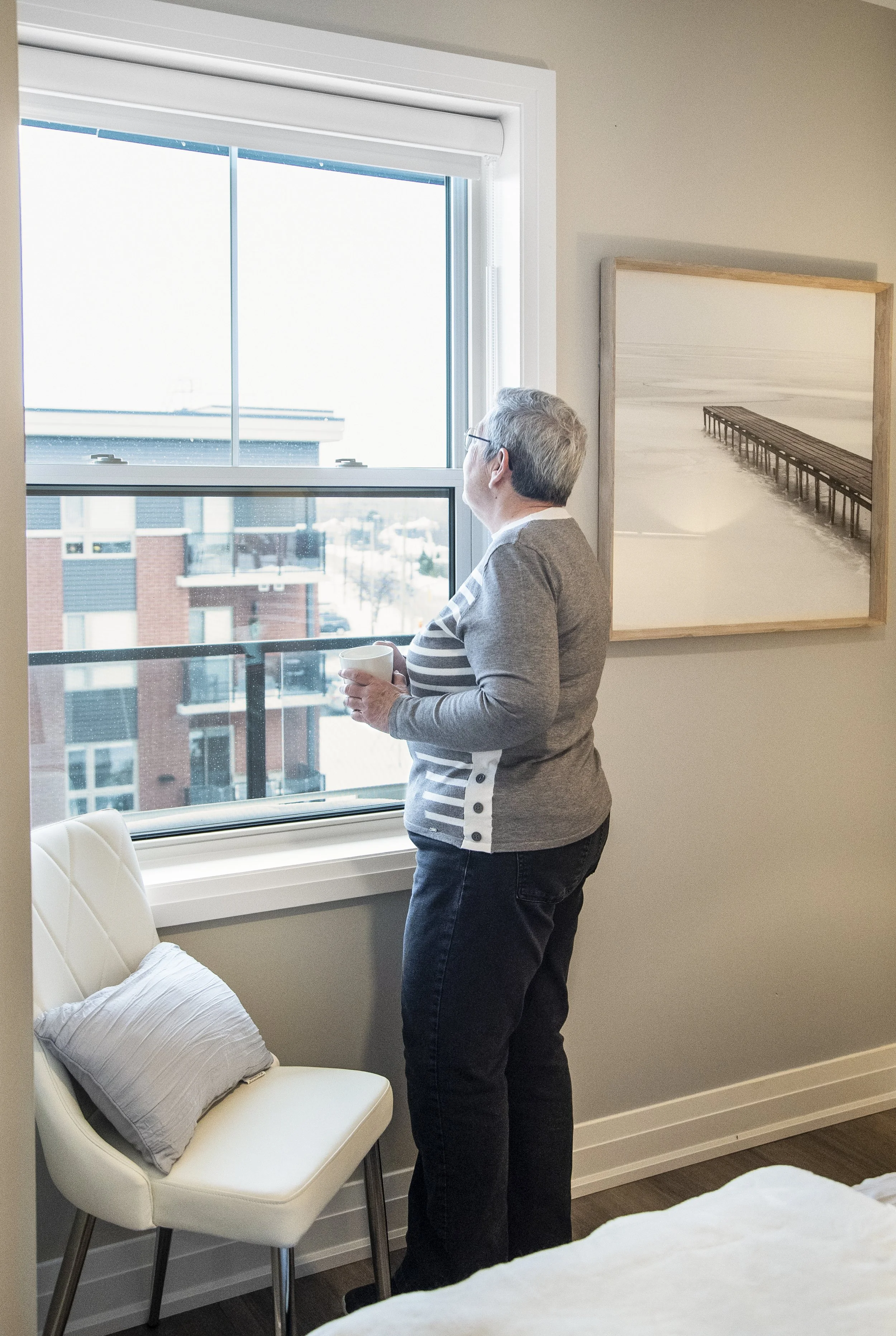 An older woman with short gray hair and glasses, wearing a gray and white striped sweater, stands by a window holding a mug, looking outside in a modern apartment with a painting on the wall and a white chair with a pillow.