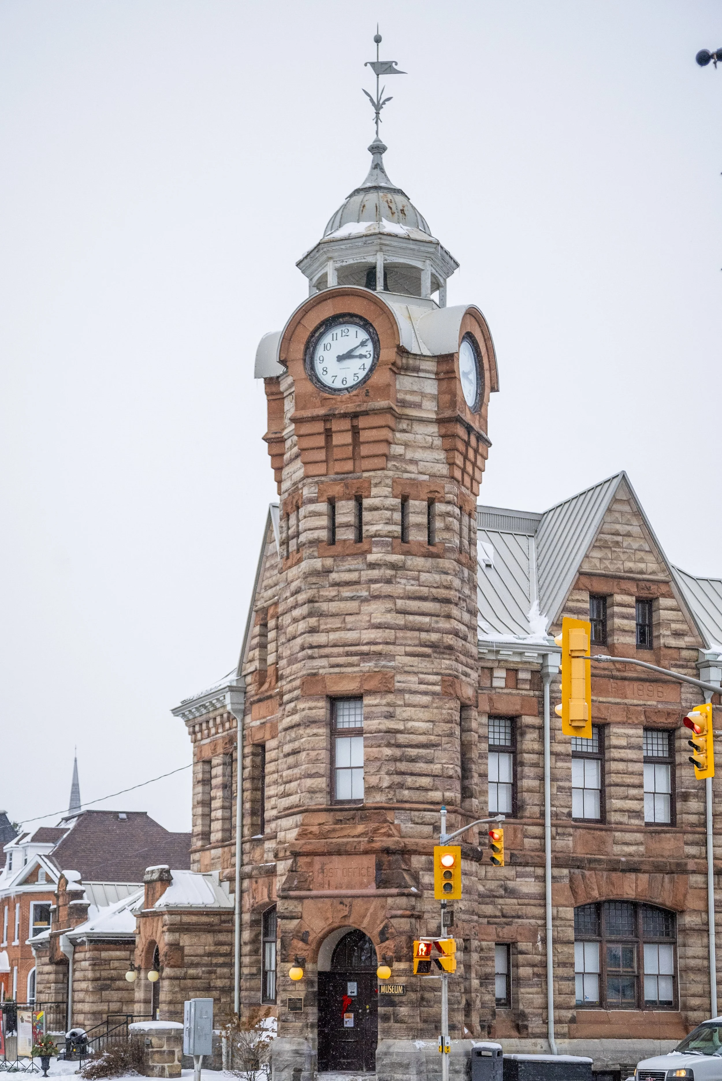 A historic brick clock tower building with a tall spire, showing the time as 10:12, in a city with snow on the ground and traffic signals in front.