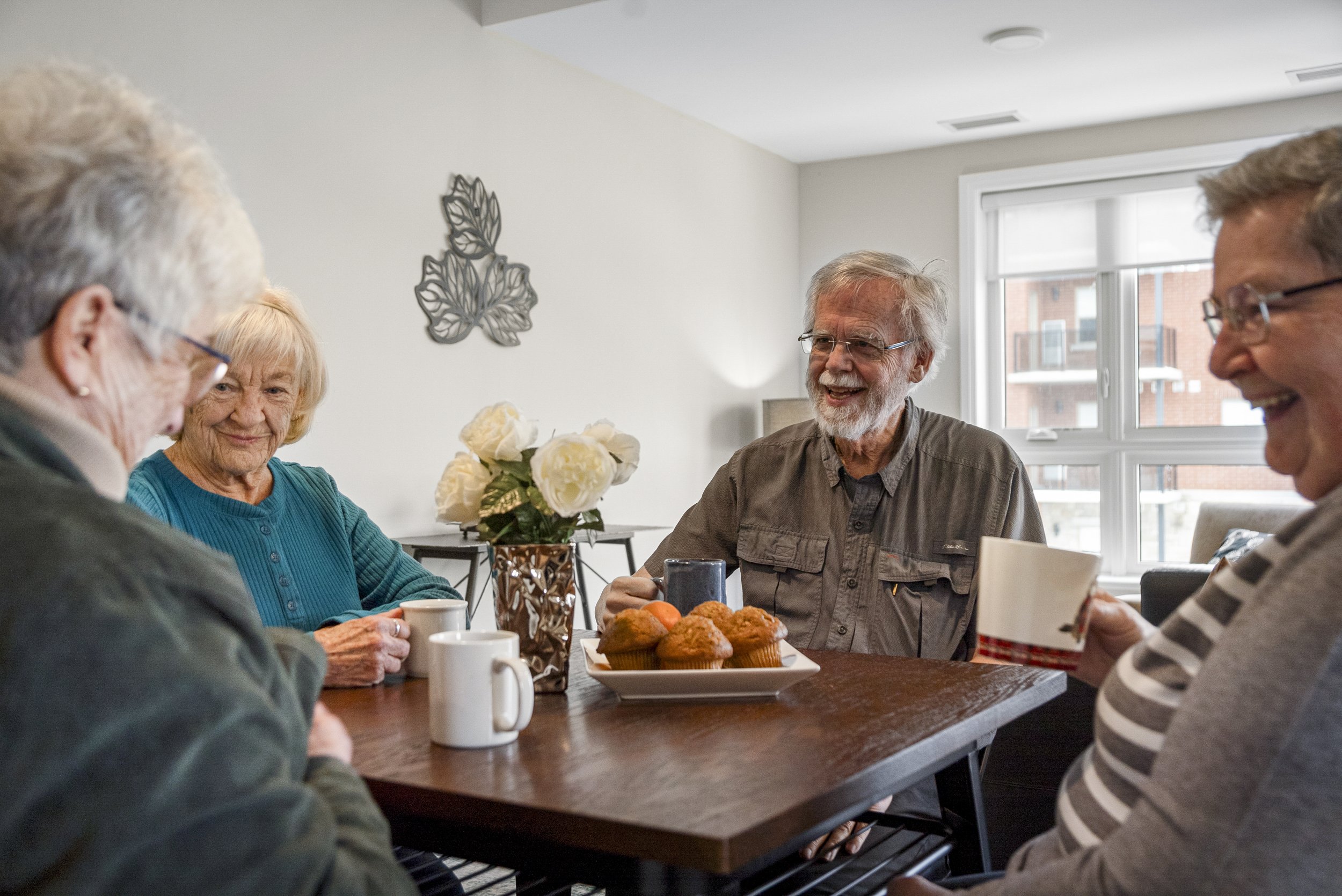Group of older adults sitting at a dining table enjoying coffee and baked goods, smiling and talking in a bright room with large windows.