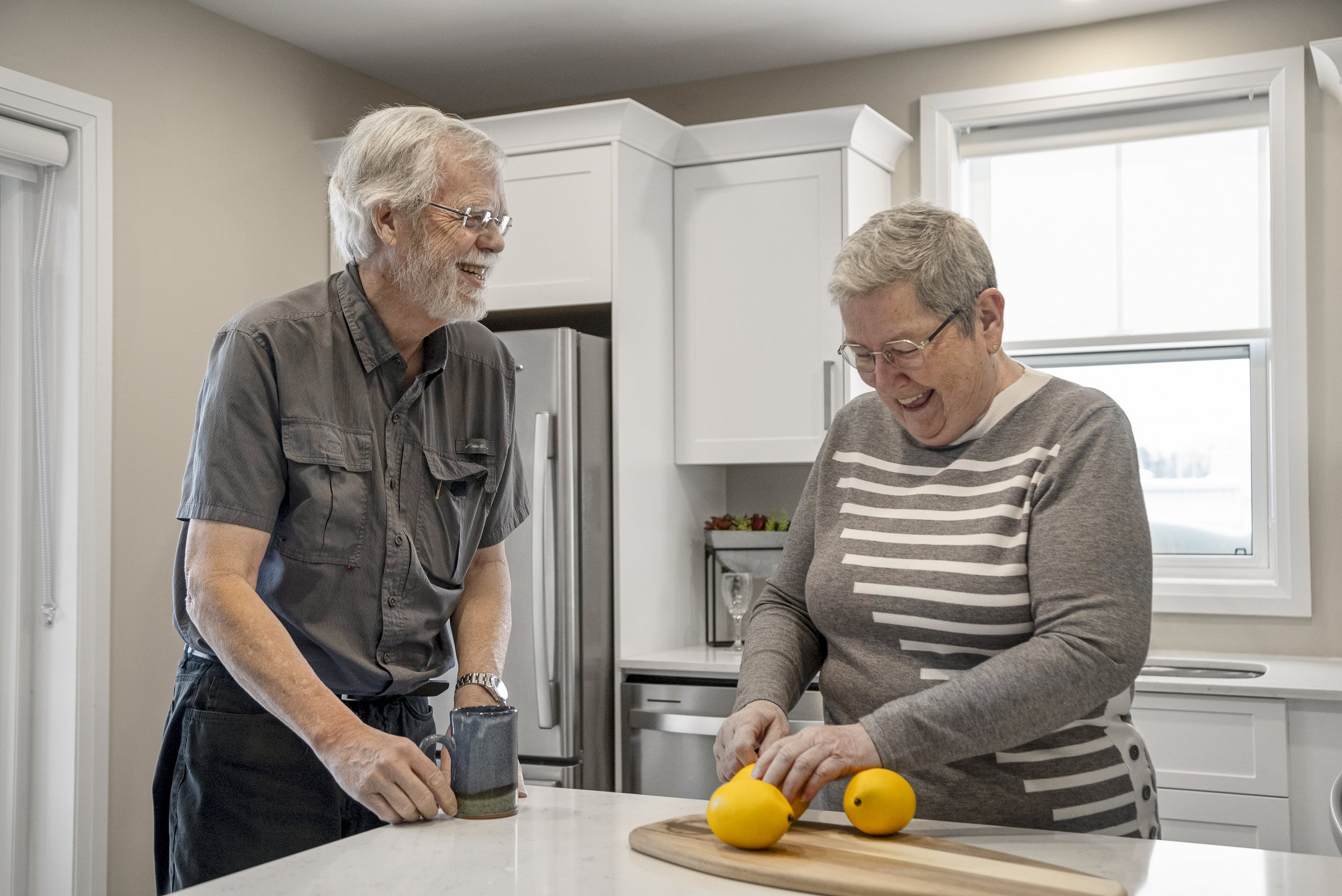 An elderly man and woman share a laugh in a kitchen. The man is holding a mug, and the woman is slicing lemons on a wooden cutting board.