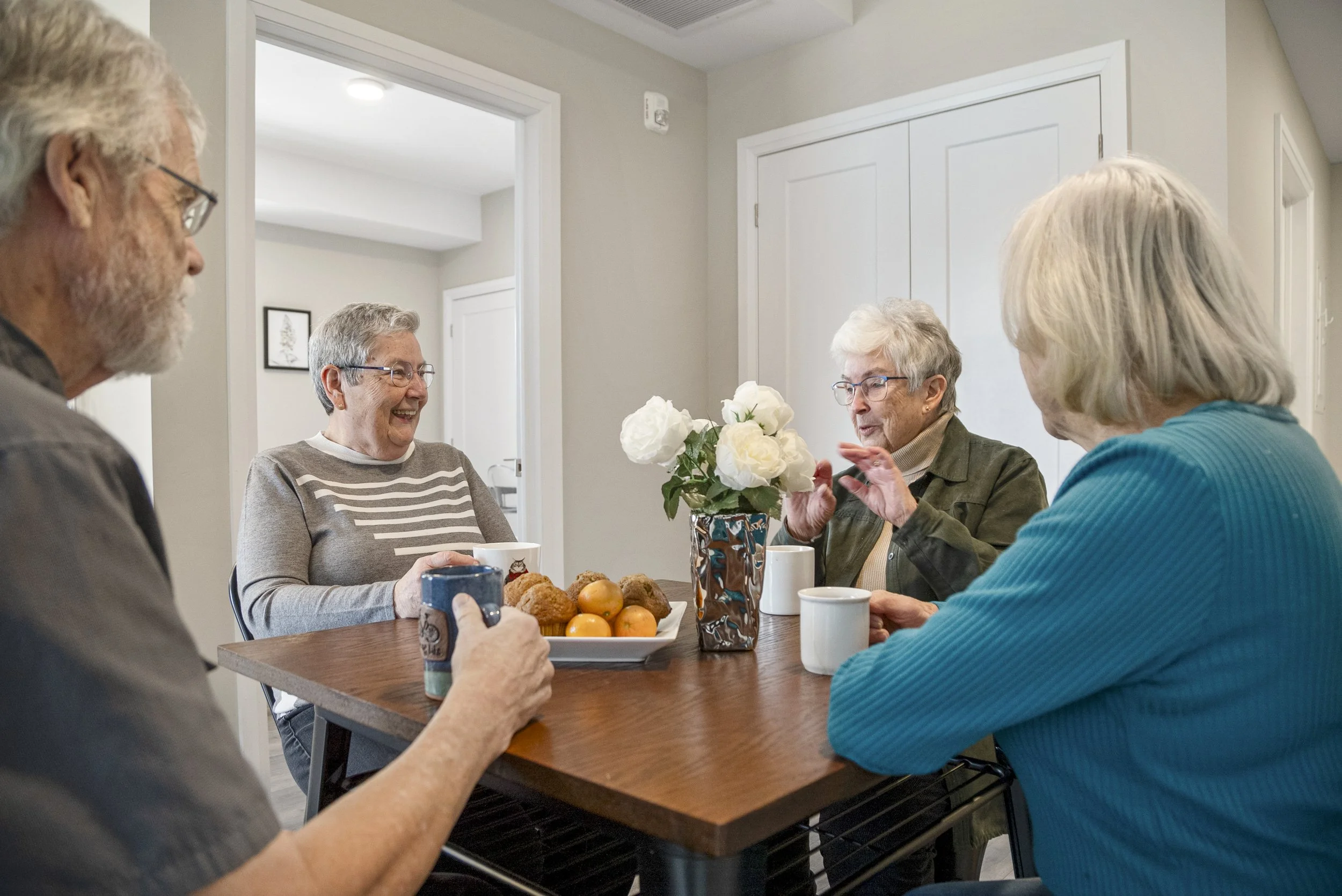 Group of five elderly people sitting around a wooden table, having a conversation, with a vase of white flowers and a tray of fruit and pastries on the table.