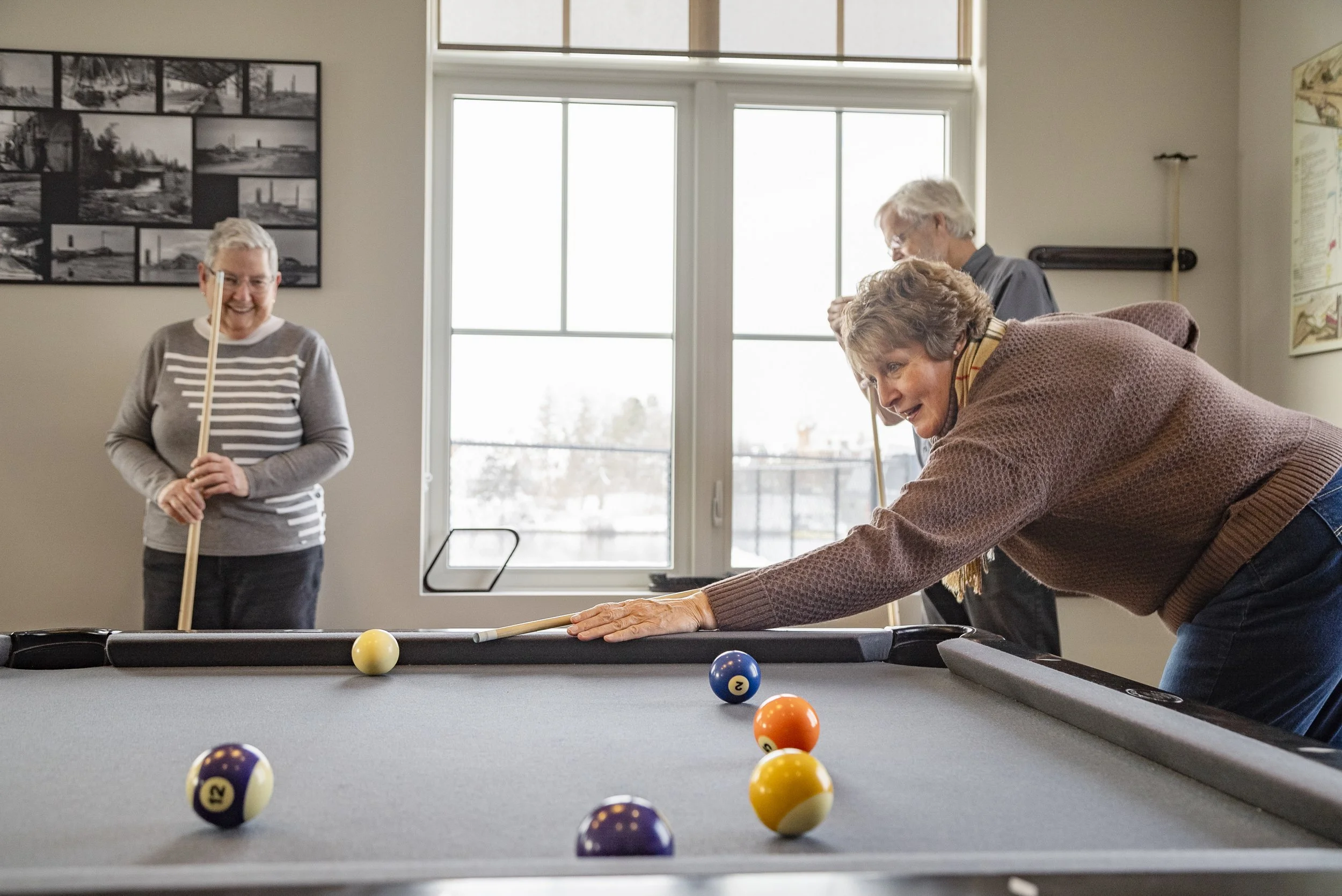 Three elderly women playing pool in a well-lit room, one leaning over to take a shot, two smiling women observing, with pool balls scattered on the table and a window providing natural light.