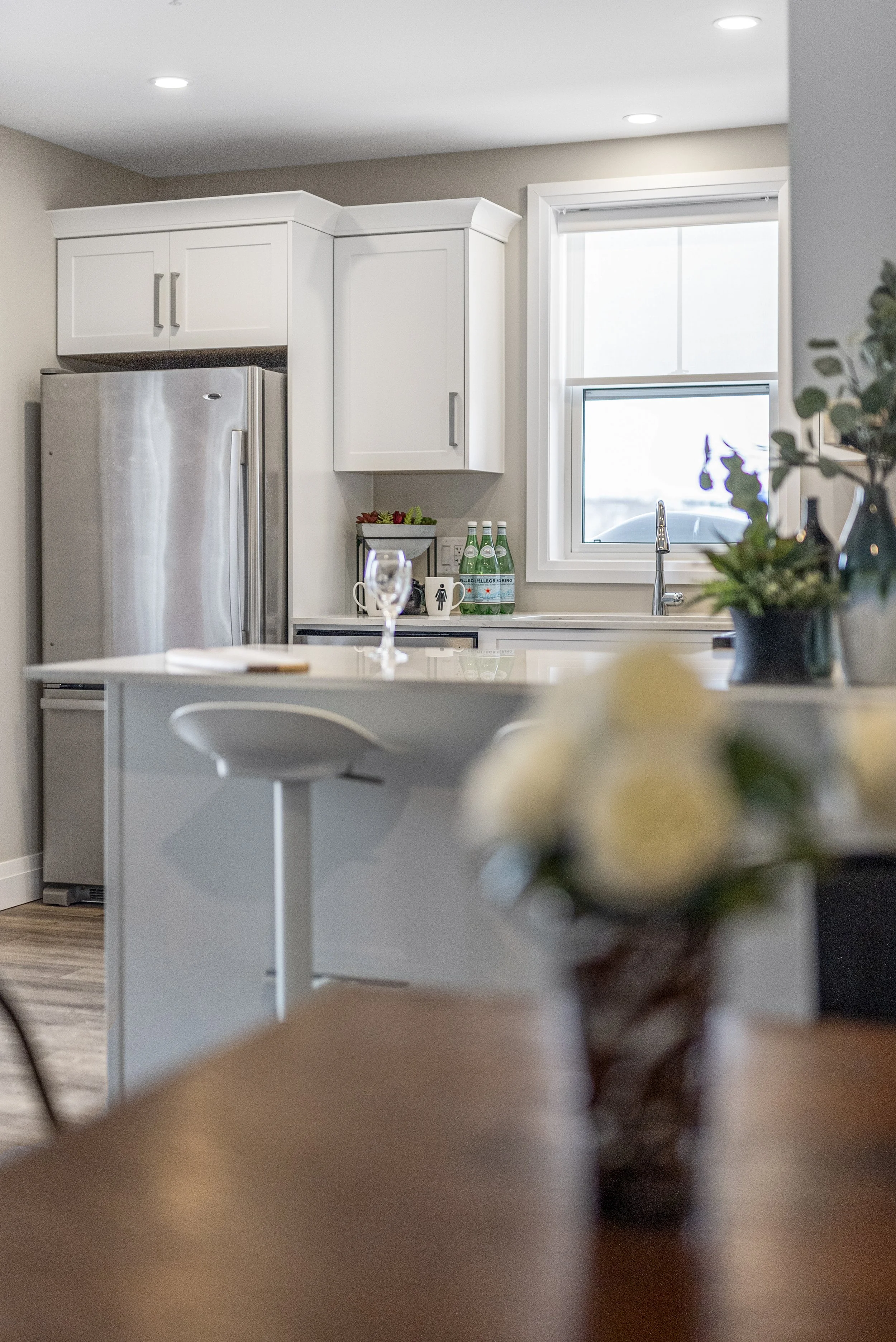 A modern kitchen with white cabinets, a stainless steel refrigerator, a window above the sink, and a kitchen island with a blurred flower arrangement in the foreground.