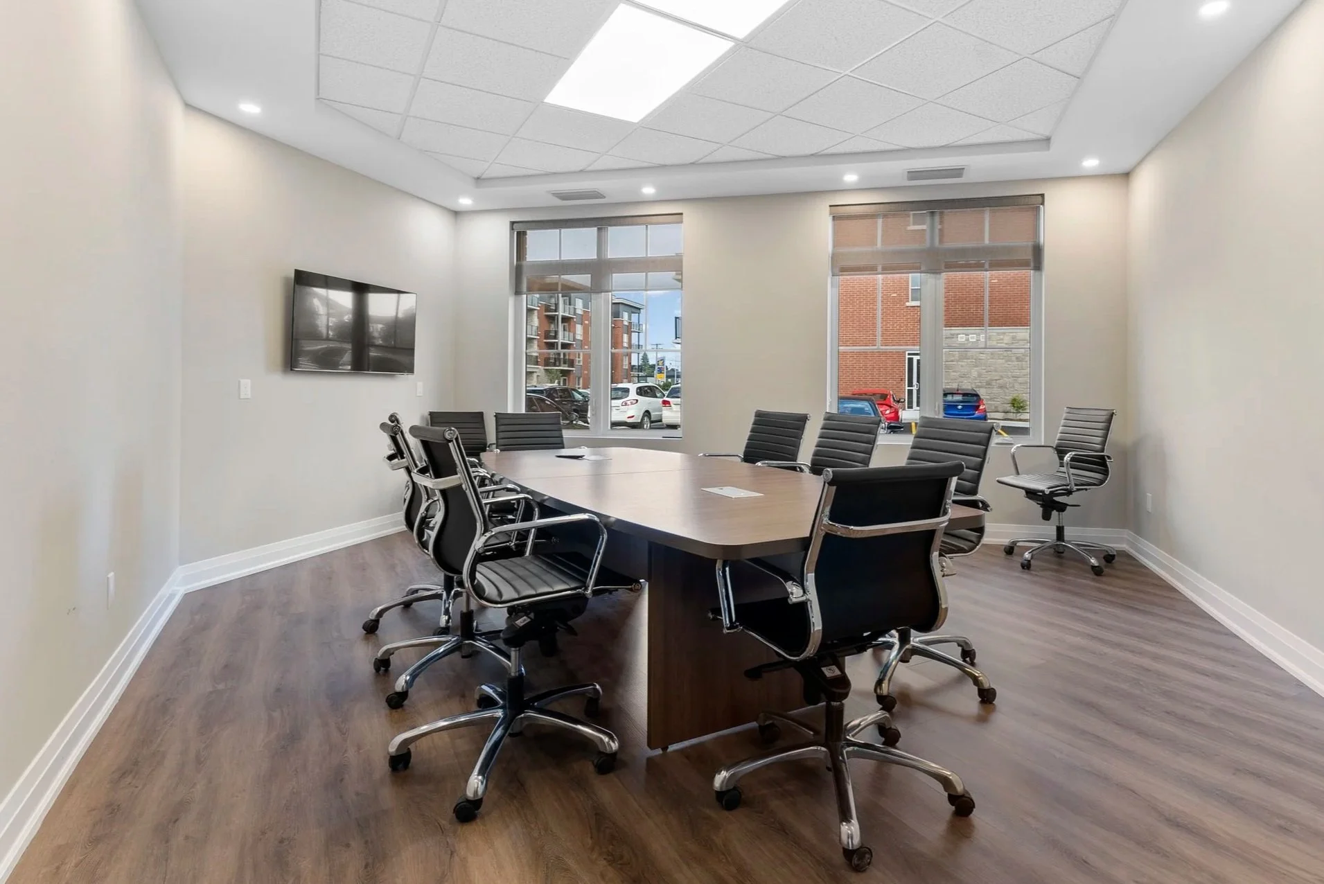 An empty conference room with a modern wooden table, black executive chairs, a wall-mounted TV, large windows showing a city street, and recessed ceiling lights.