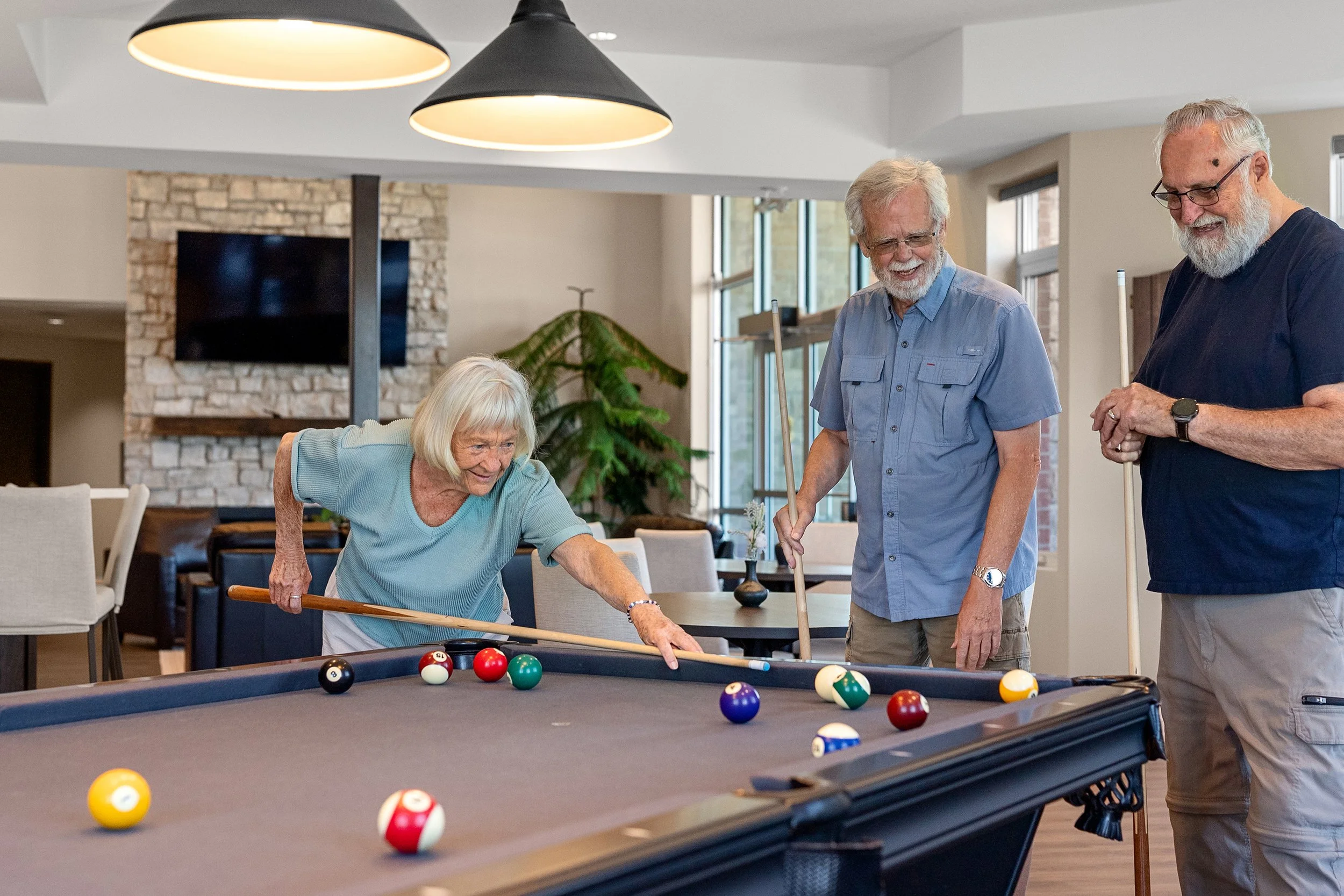 A group of older adults smiling and playing pool around a billiards table in a well-lit lounge.