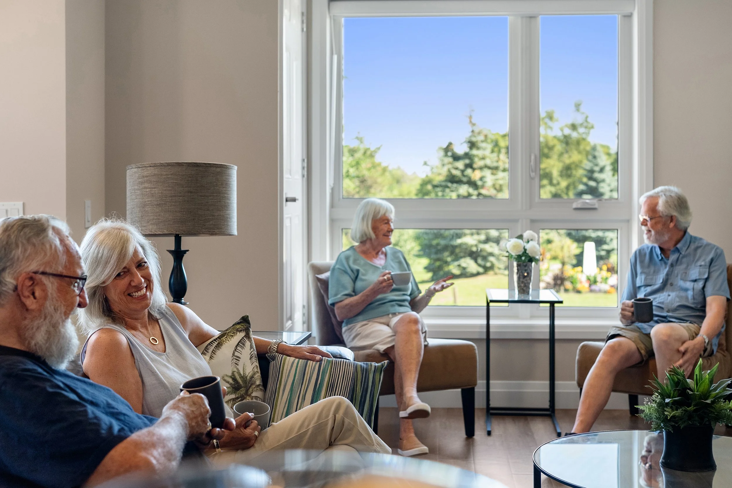 Four older adults sitting together in a living room near a large window, enjoying coffee and conversation.